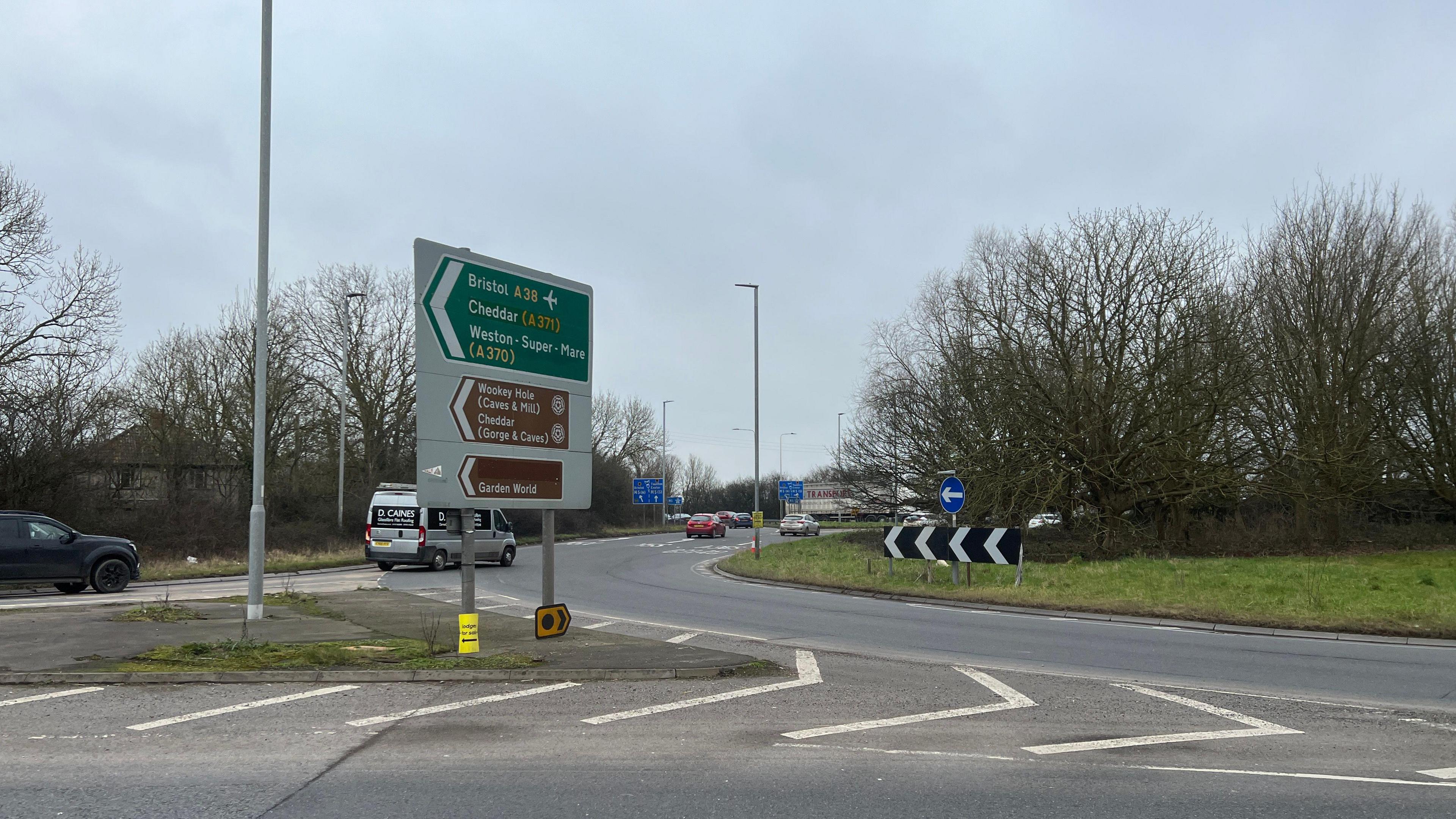 A roundabout with several lanes. There's a green sign pointing north saying A38 to Cheddar and Bristol Airport. There's a brown sign saying Cheddar Gorge and Wookey Hole. There's a view cars in shot entering the roundabout.