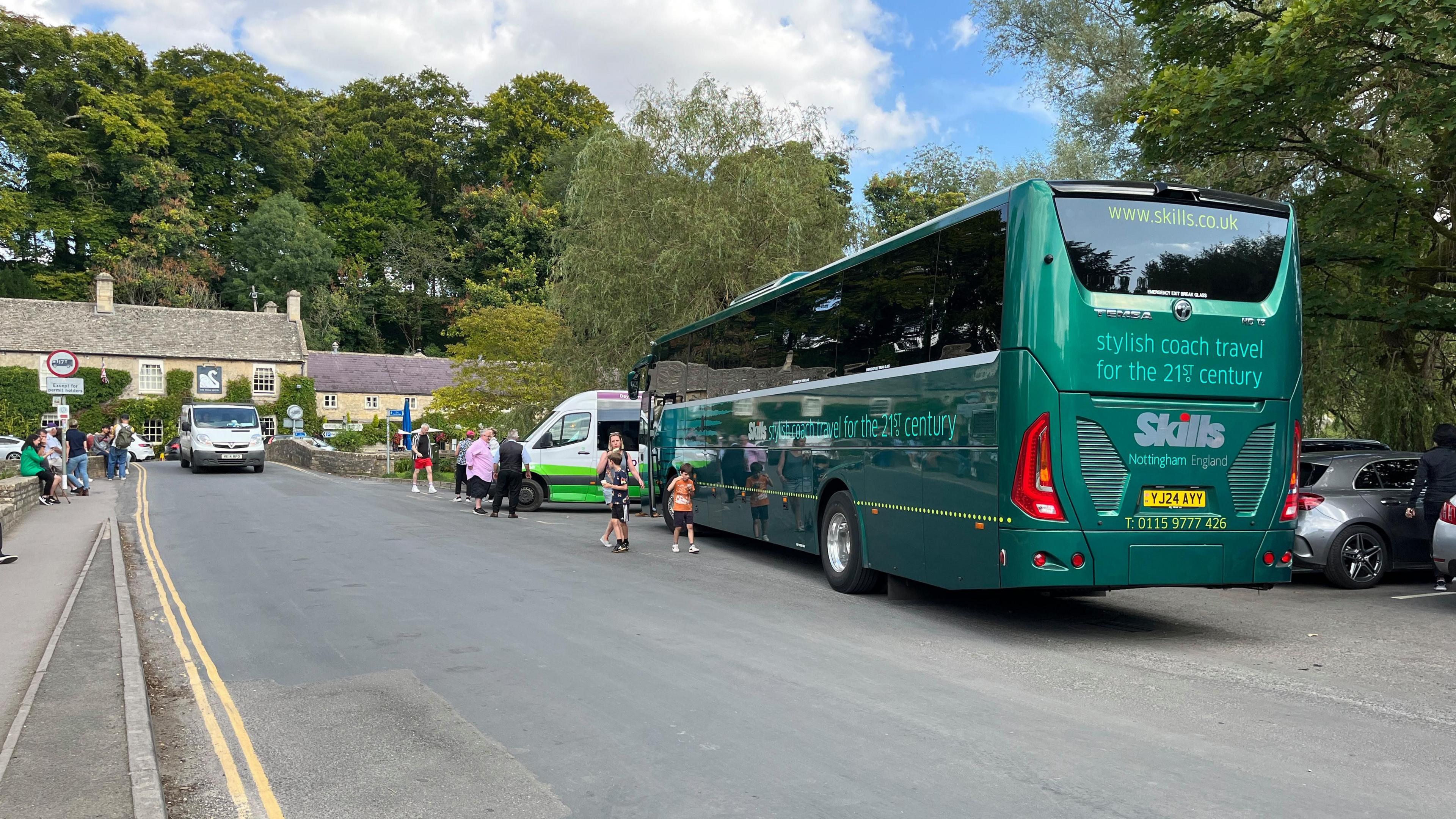 A large green coach is parked on a narrow road in Bibury. In the distance a van drives the opposite direction. People are walking around.