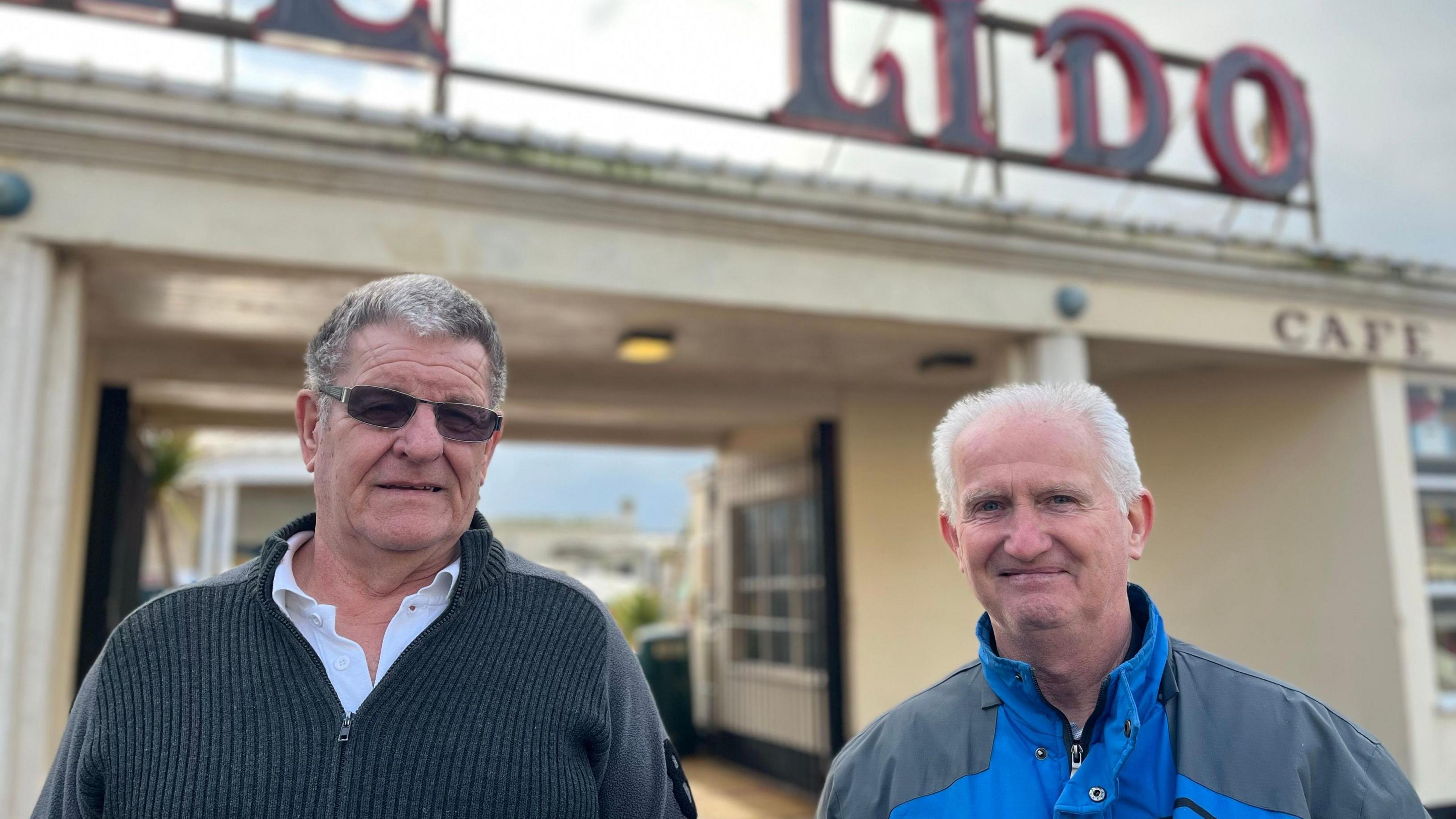 Richard White wearing sunglasses and a grey jumper, and Peter Planner wearing a blue coat both standing looking at the camera outside the front of the lido which has a large red sign which reads The Lido