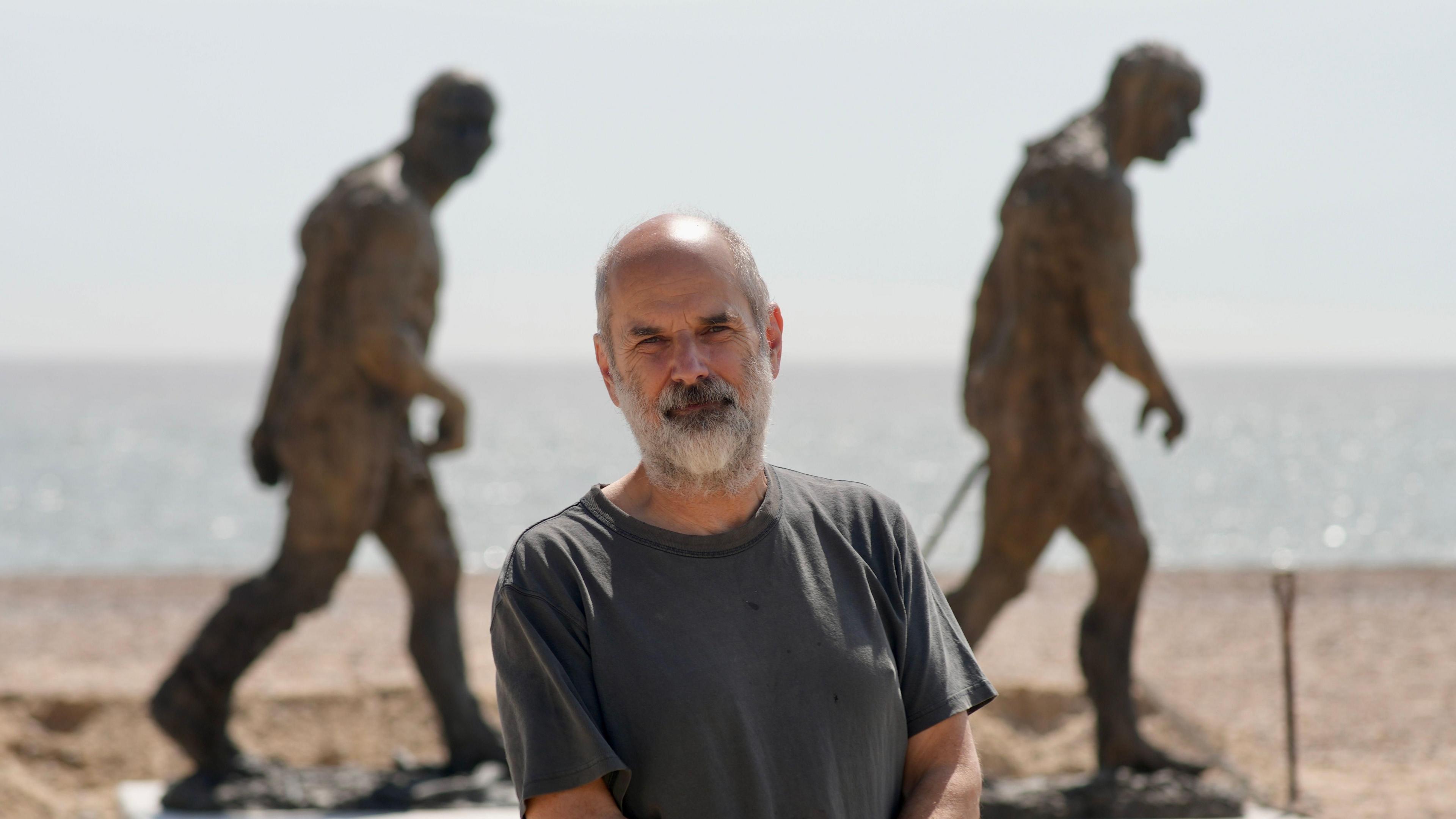 Laurence Edwards with short grey hair and beard wearing a grey t-shirt on a beach with two bronze men in a walking pose behind him.  The sea is visible behind the sculpture.