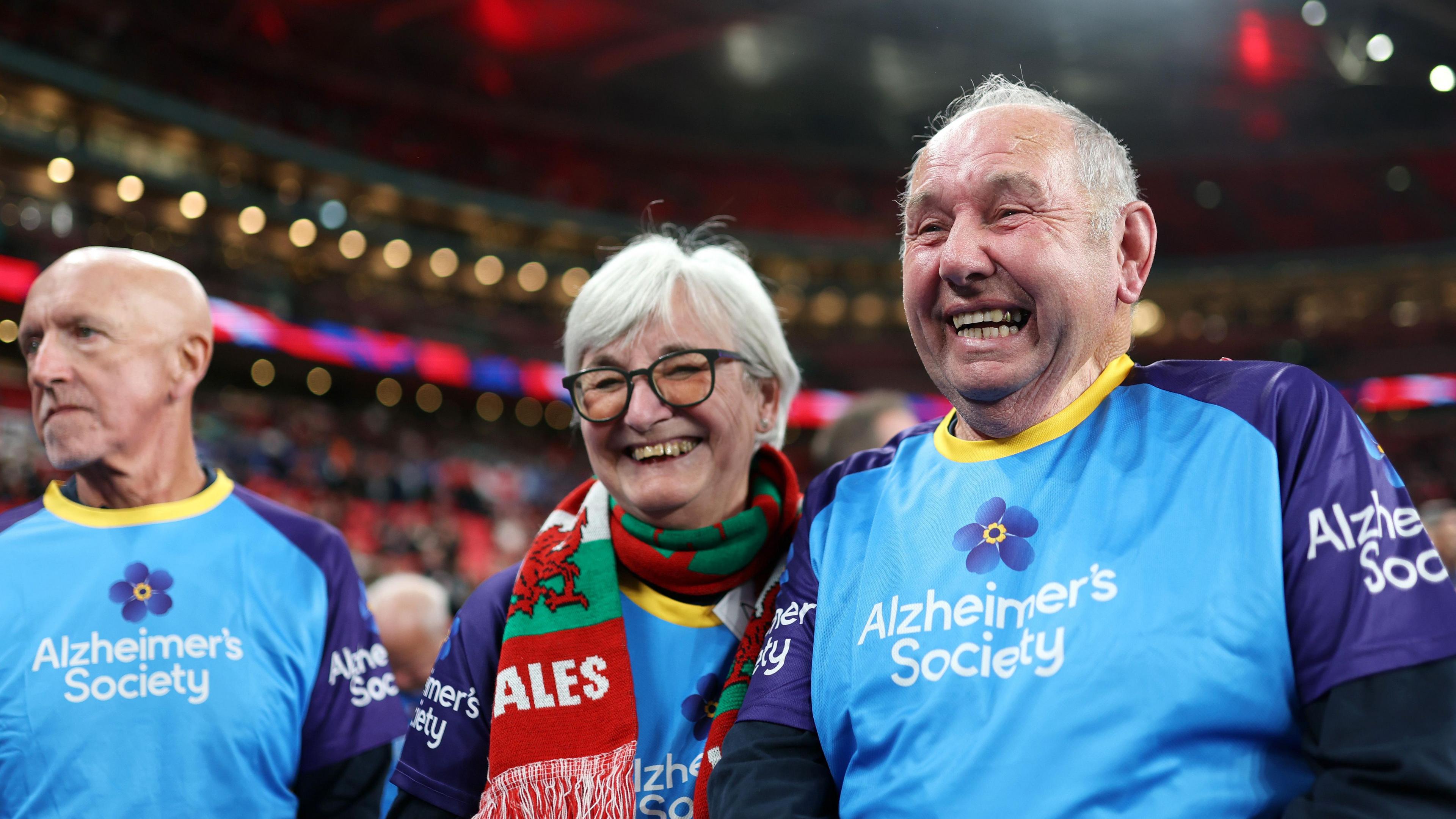Mascots are seen pitchside during an Alzheimer's Society Partner Activity during the Men's International Friendly match between England and Wales at Wembley Stadium on October 09, 2025 in London. A man and a woman grin at the camera, they are wearing the blue tops that say Alzheimer's Society. The woman, who has short grey hair, is wearing a Wales scarf and glasses.