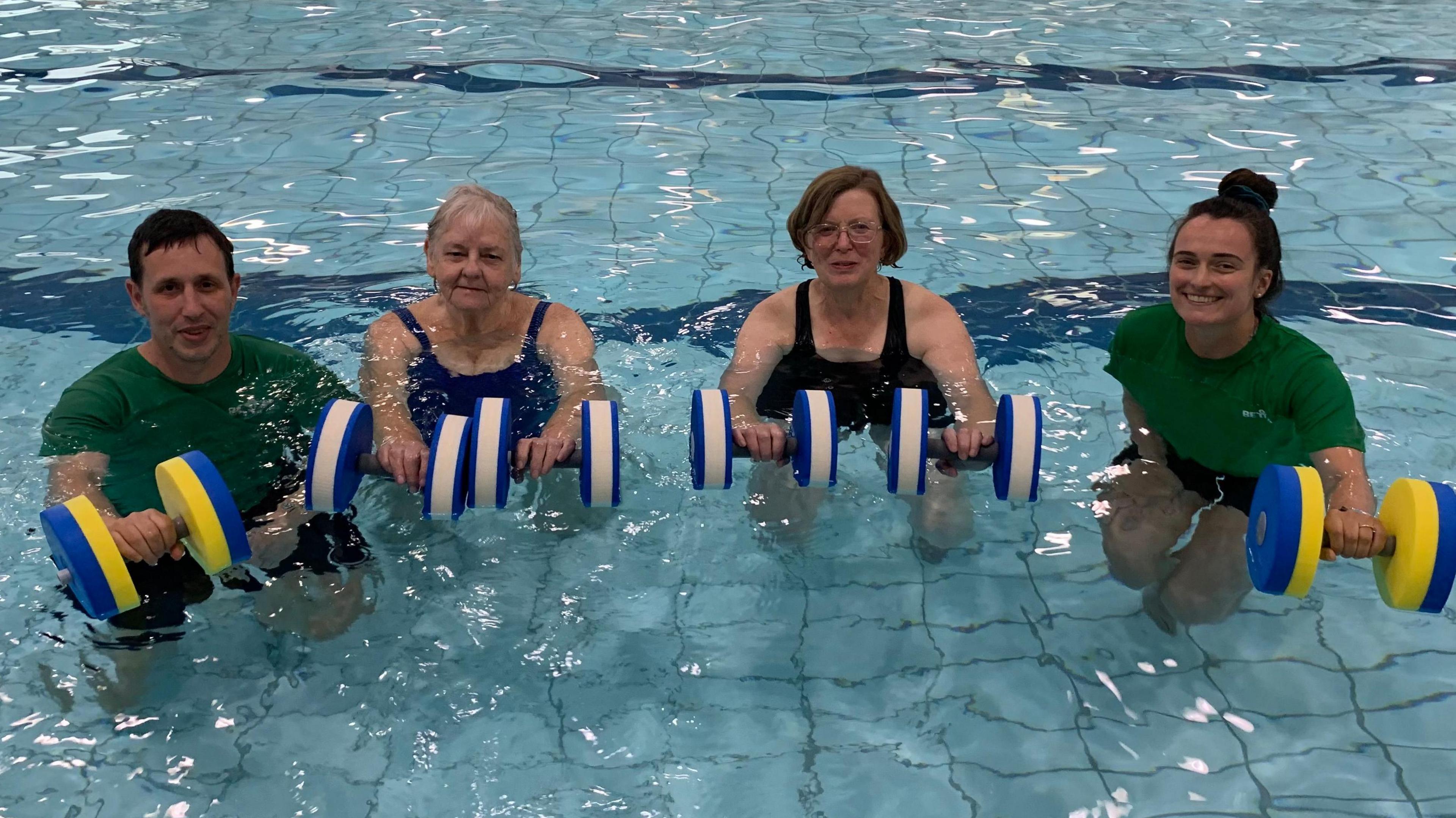 Ms Vipond Houghton in Kendal pool with another member of staff, both wearing green polo shirts. Between them are two members of her class, wearing swimming costumes. They are are all holding floating dumbbells, some blue and yellow, others blue and pink.