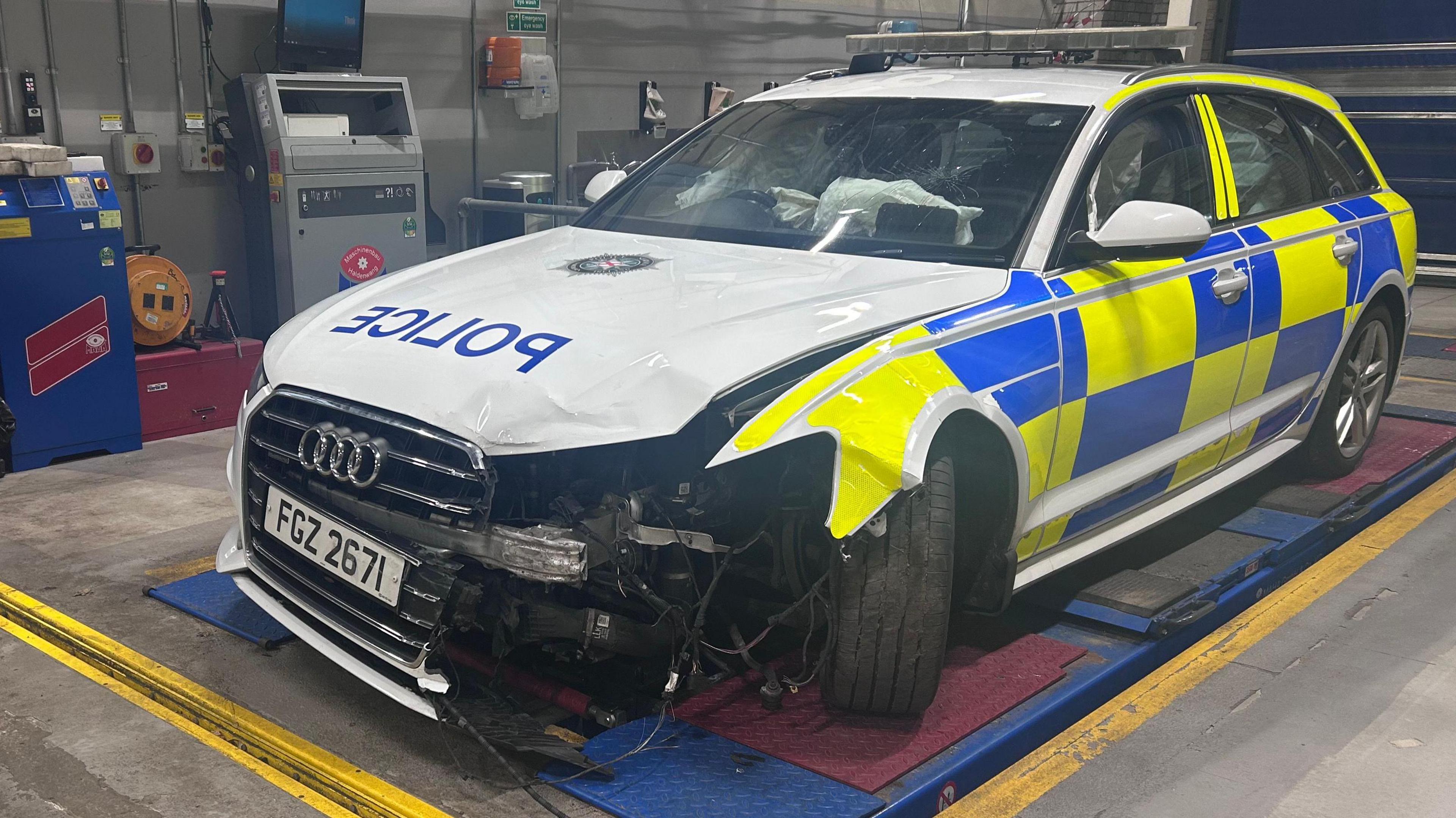 A damged police Audi sits in a garage. It is blue and yellow police colours, with a white bonnet. Their is a chunk taken out of the front left of the car, inbetween the grill and the left wheel. Inside the car airbags have been deployed. 