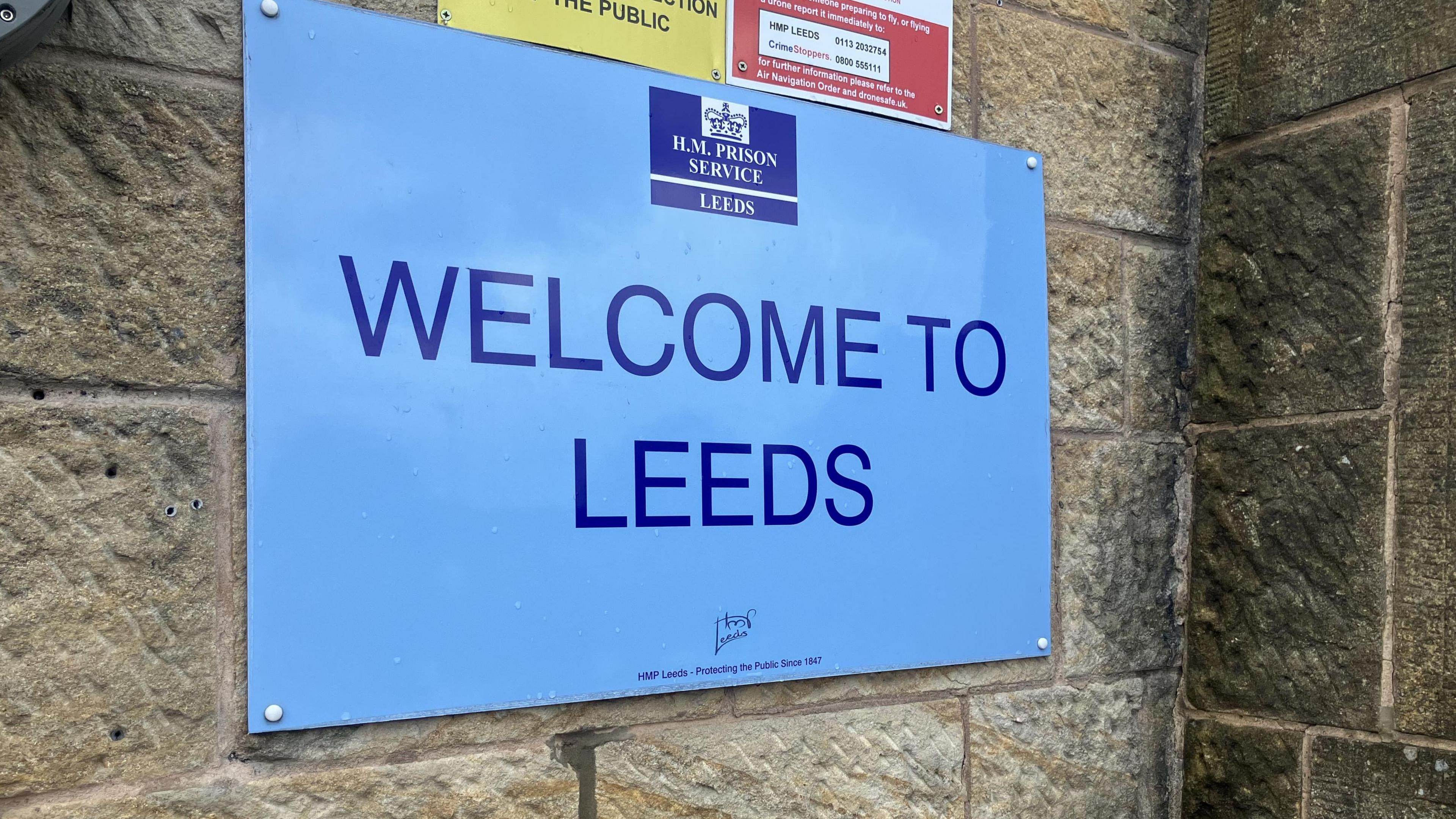 A blue sign on a wall saying 'Welcome to Leeds'. The logo of HM Prison Service is at the top.