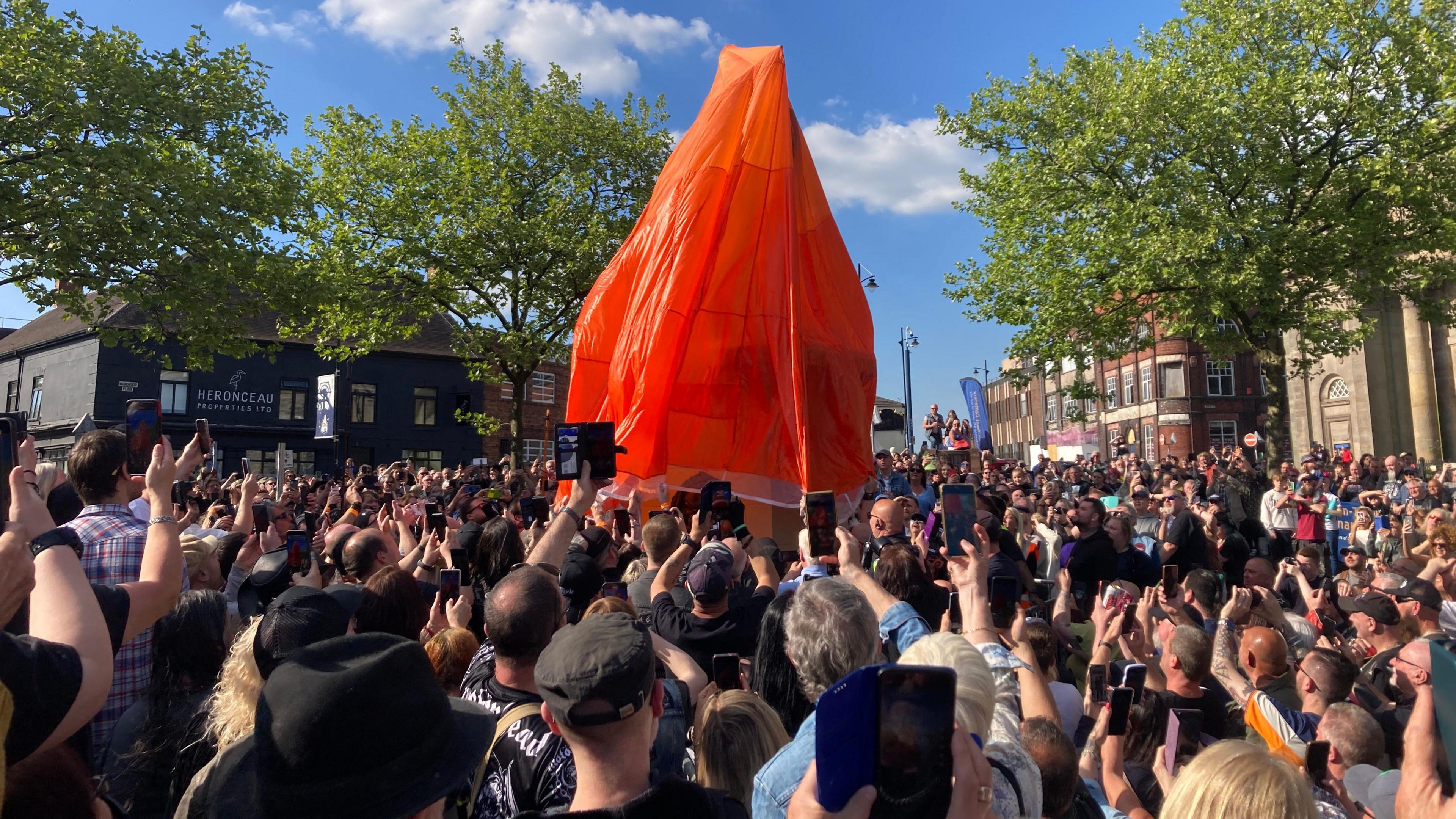 A view from the back of a large crowd standing outside in a square with phones raised ready to take a picture. A red sheet covers a statue at the centre of the photo. Buildings surround the square and the sky is blue.