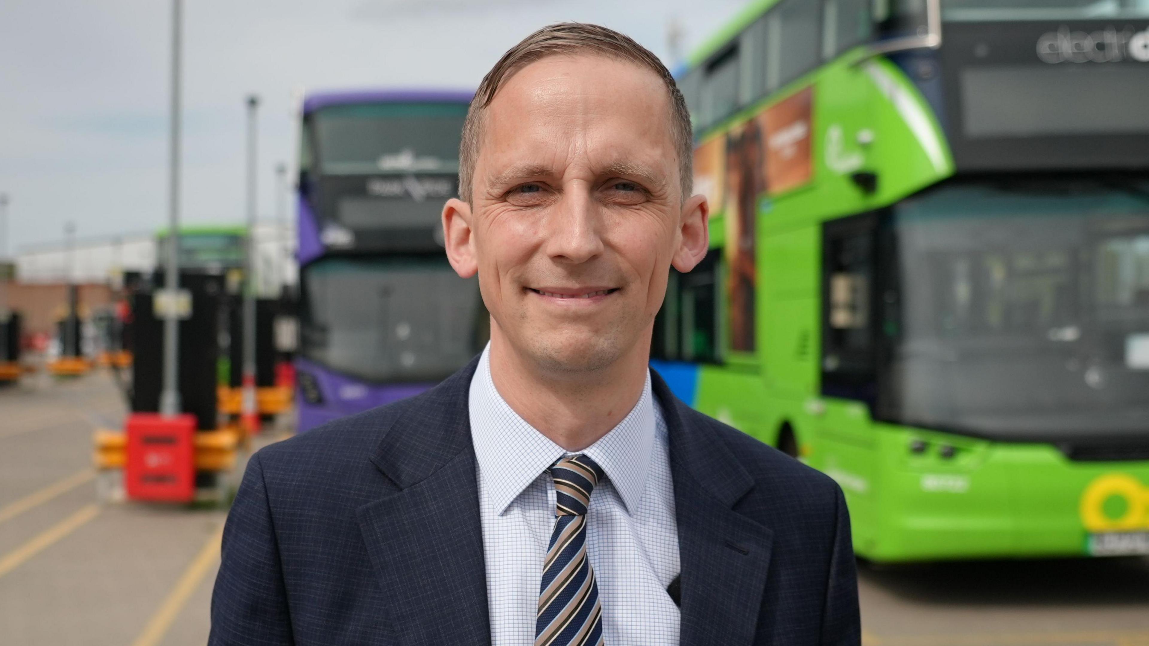 Mr Marion has short light brown hair, and wears a blue suit and tie. Two buses are behind him.