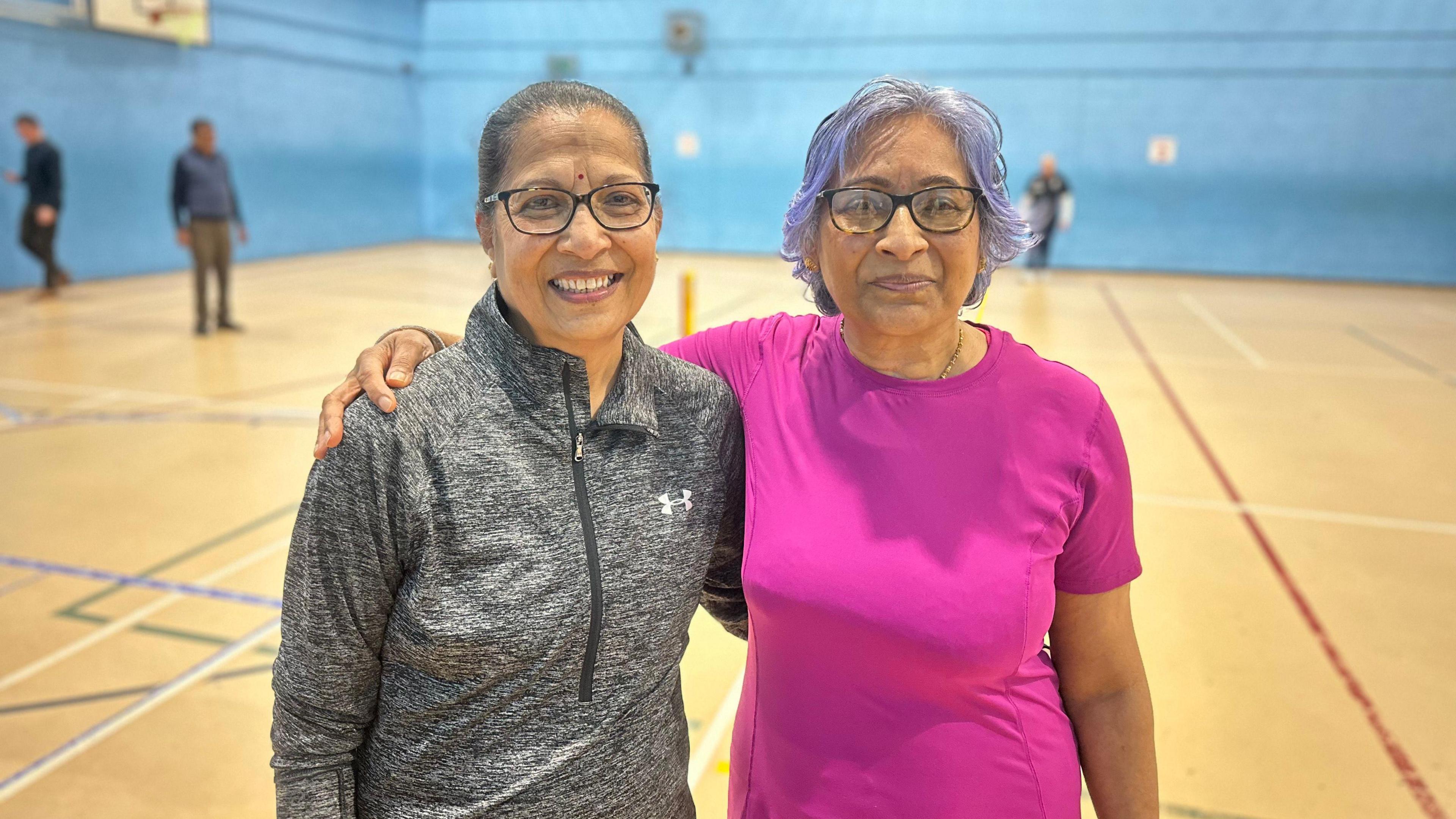 Two women are posing with their arms around each other in a sports hall, and there are people in the distance behind them. One has dark hair tied back and is wearing a grey top, and the other has grey hair and is in a pink top.