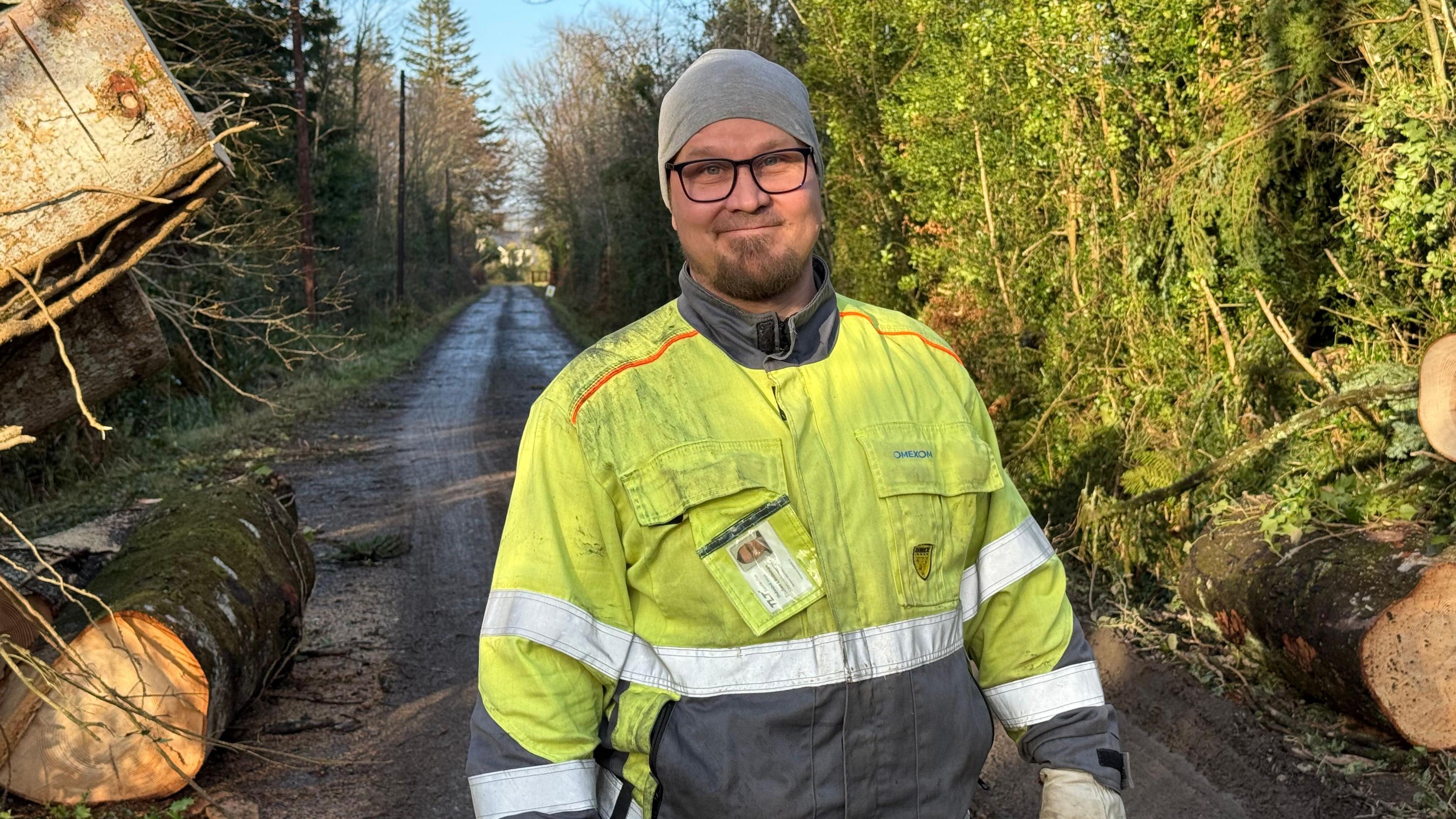 Staffan Lindeman. He is wearing a grey beanie hat, black glasses, a high viz jacket and gloves. He is standing on a single lane road with trees on either side. Cut trunks are on either side of him.