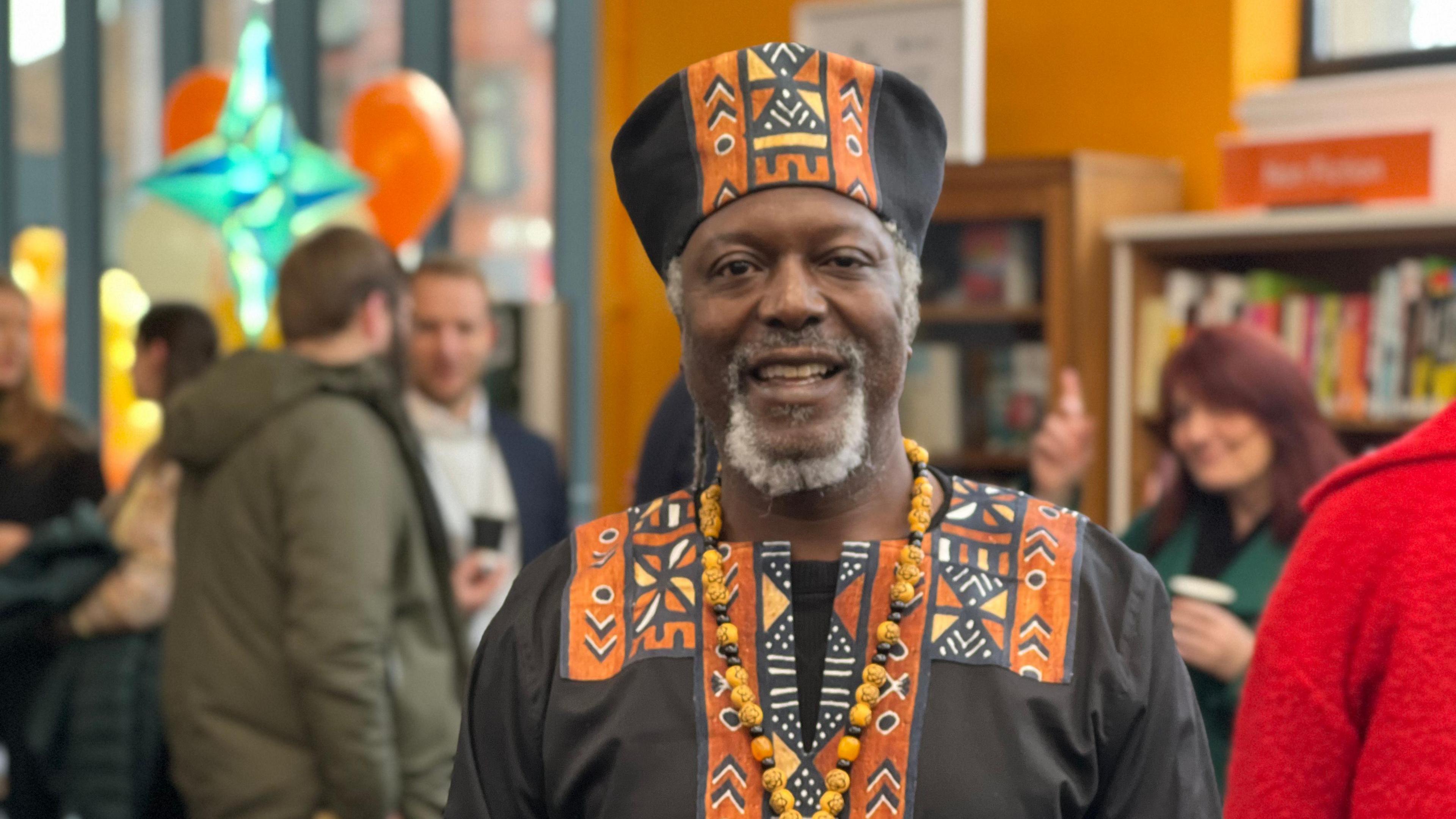 Poet Levi Tafari, who is wearing a black traditional African style gown and hat with an orange pattern, and a necklace with chunky black and orange beads, smiles at the camera