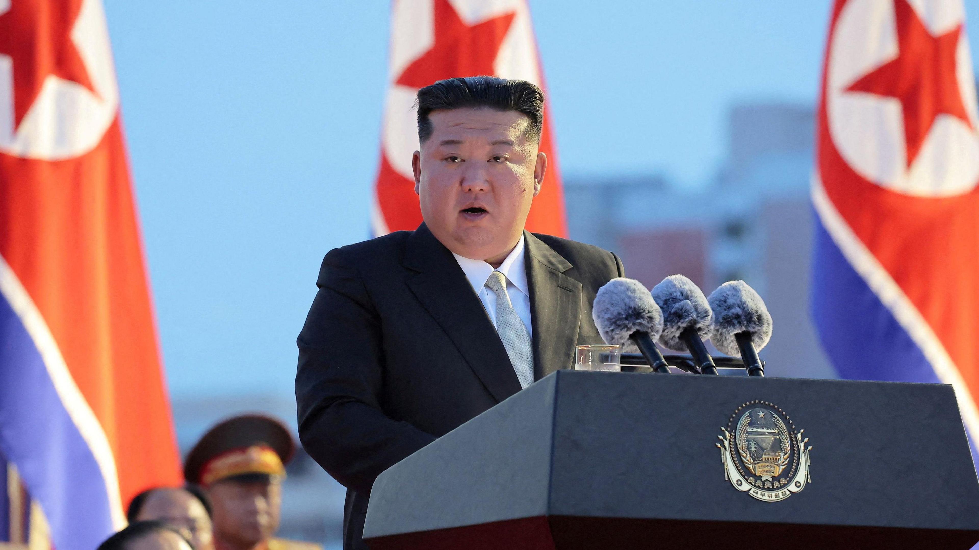 A man in a suit and tie addresses from a podium with North Korean flags in the background