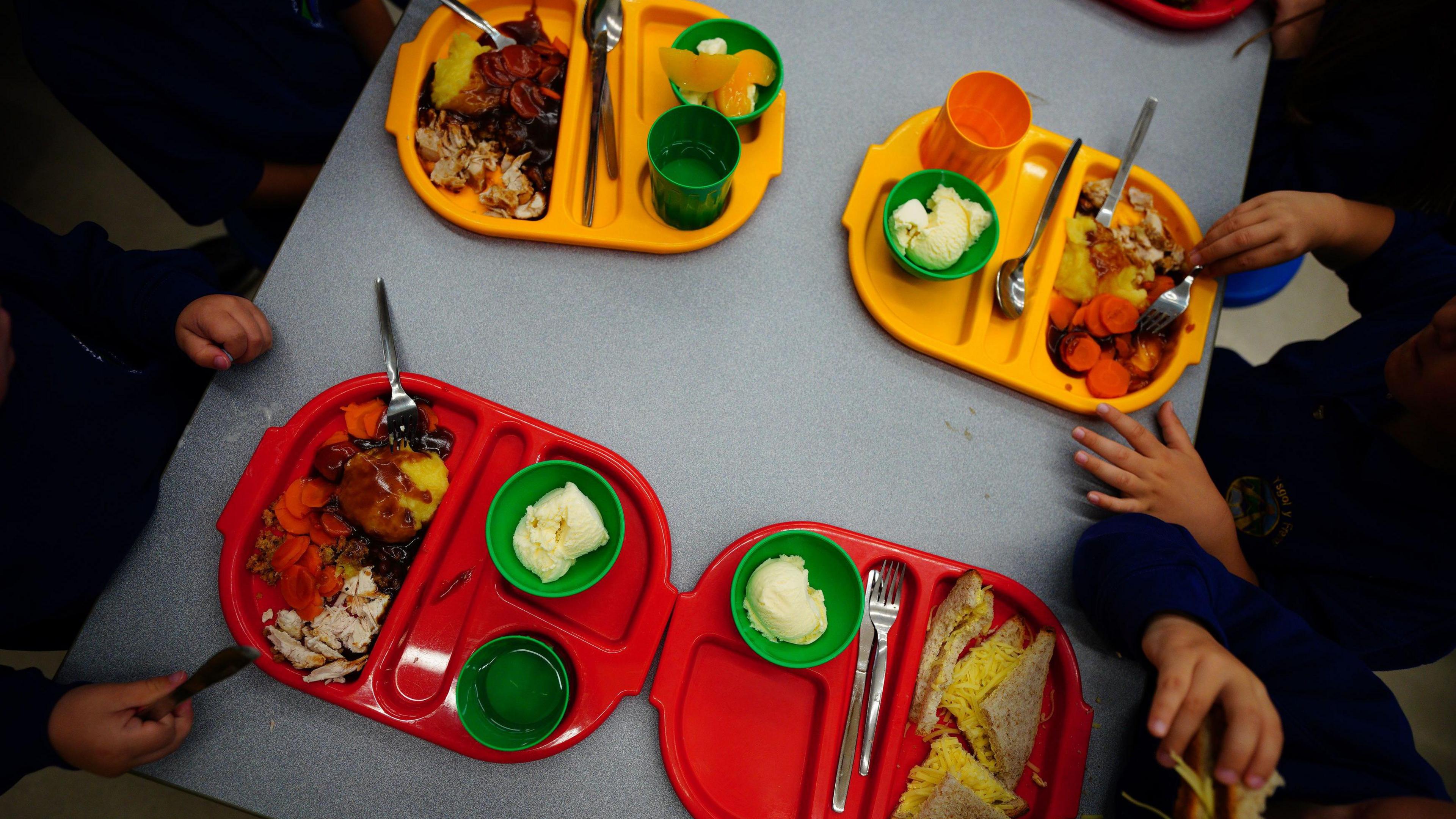 Two red and two yellow school dinner trays on a table, shown from above. The hands of the children can be seen as they tuck into their meals.