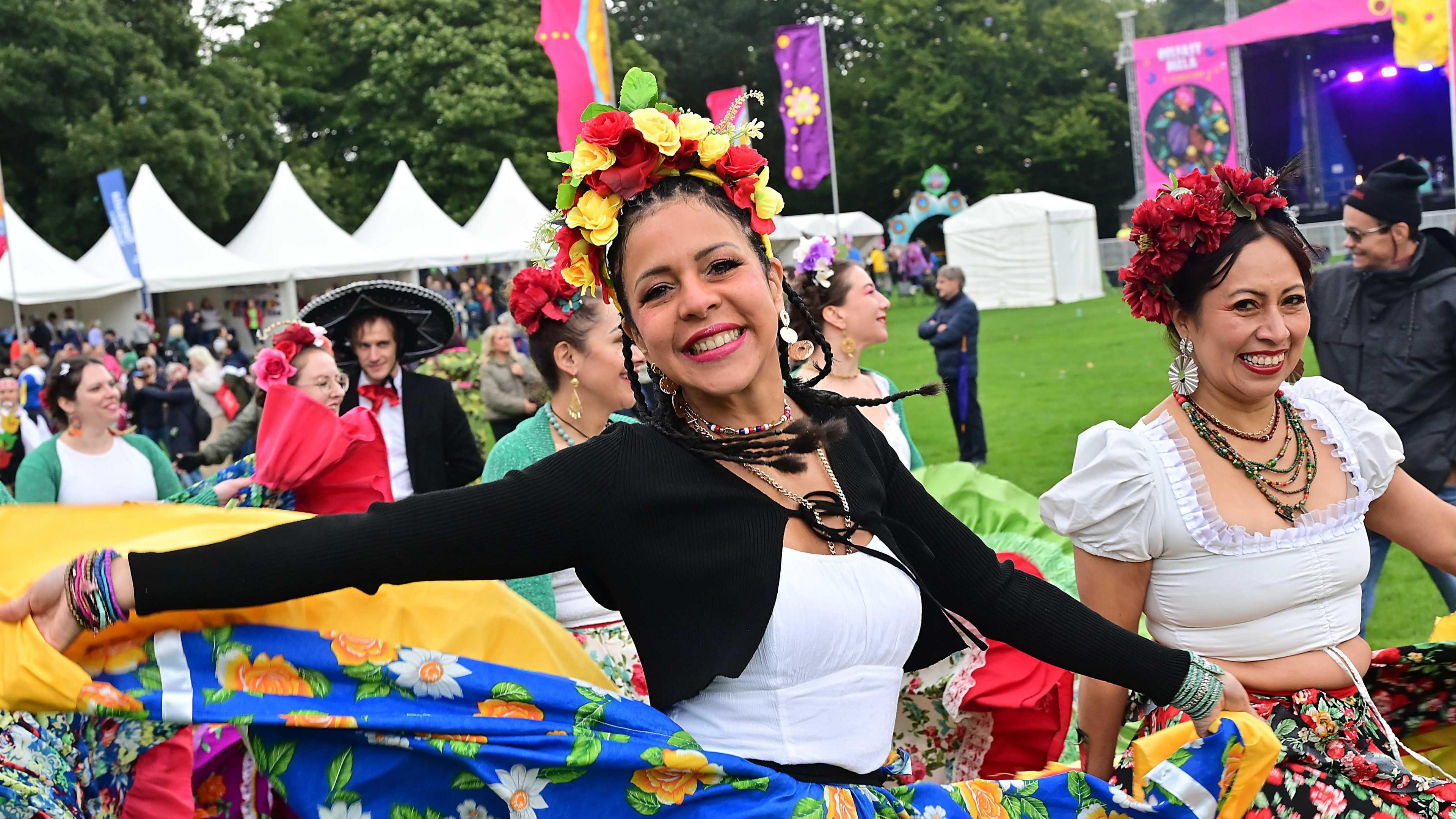 Women in flowy skirts with colourful flower patterns, dance on green grass festival grounds. They are also wearing yellow and red flowered headbands. 