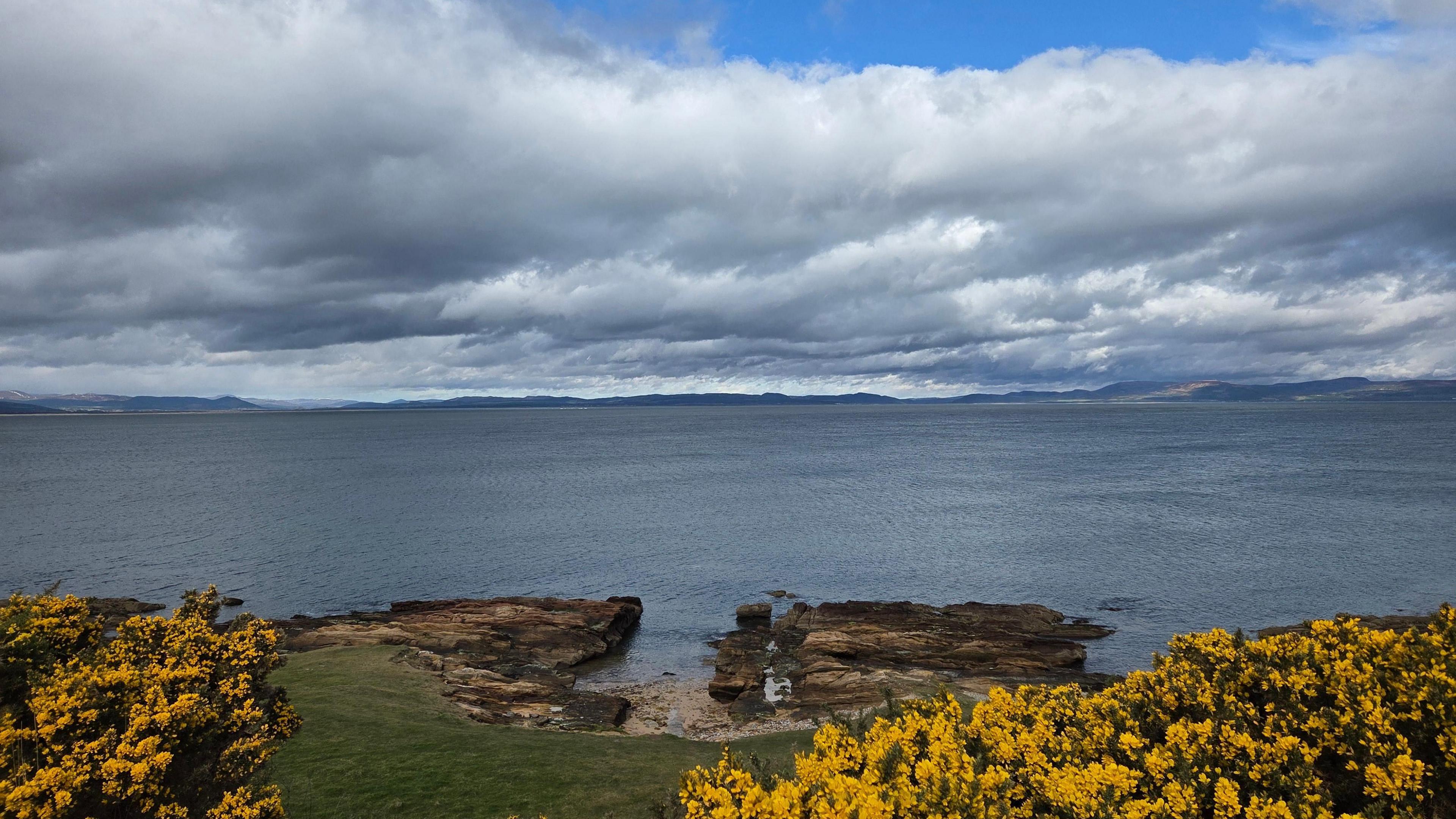 Mainly cloudy sky above the coast with rocks and yellow flowers lining the beach