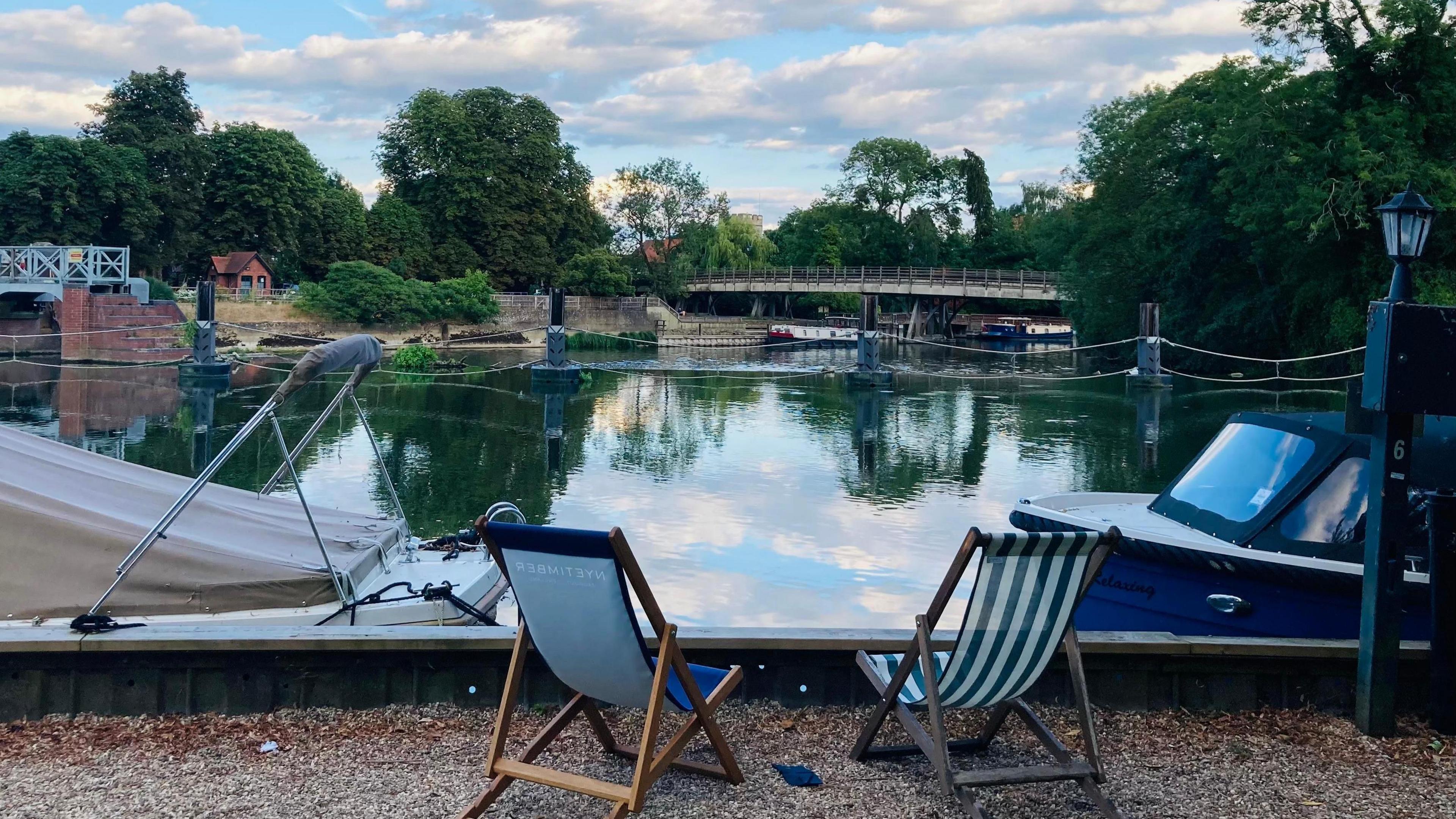 Two empty deck chairs are in the centre of the shot, facing out onto a river with two boats tied up in front of them, and a bridge in the background.