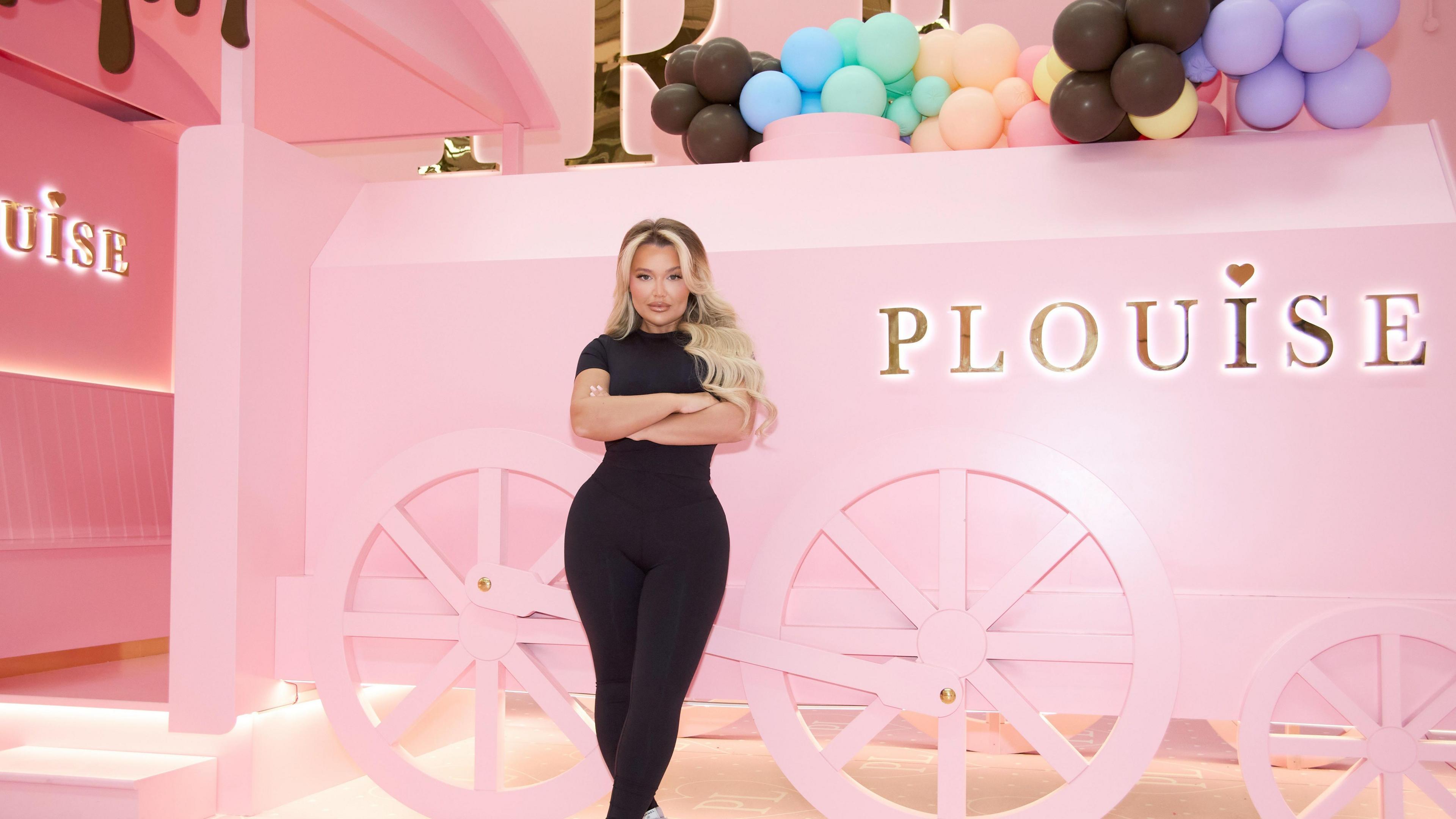 Paige wears an all black outfit, has long wanded blonde hair and is stood in front of a P.Louise model carriage with her arms folded.