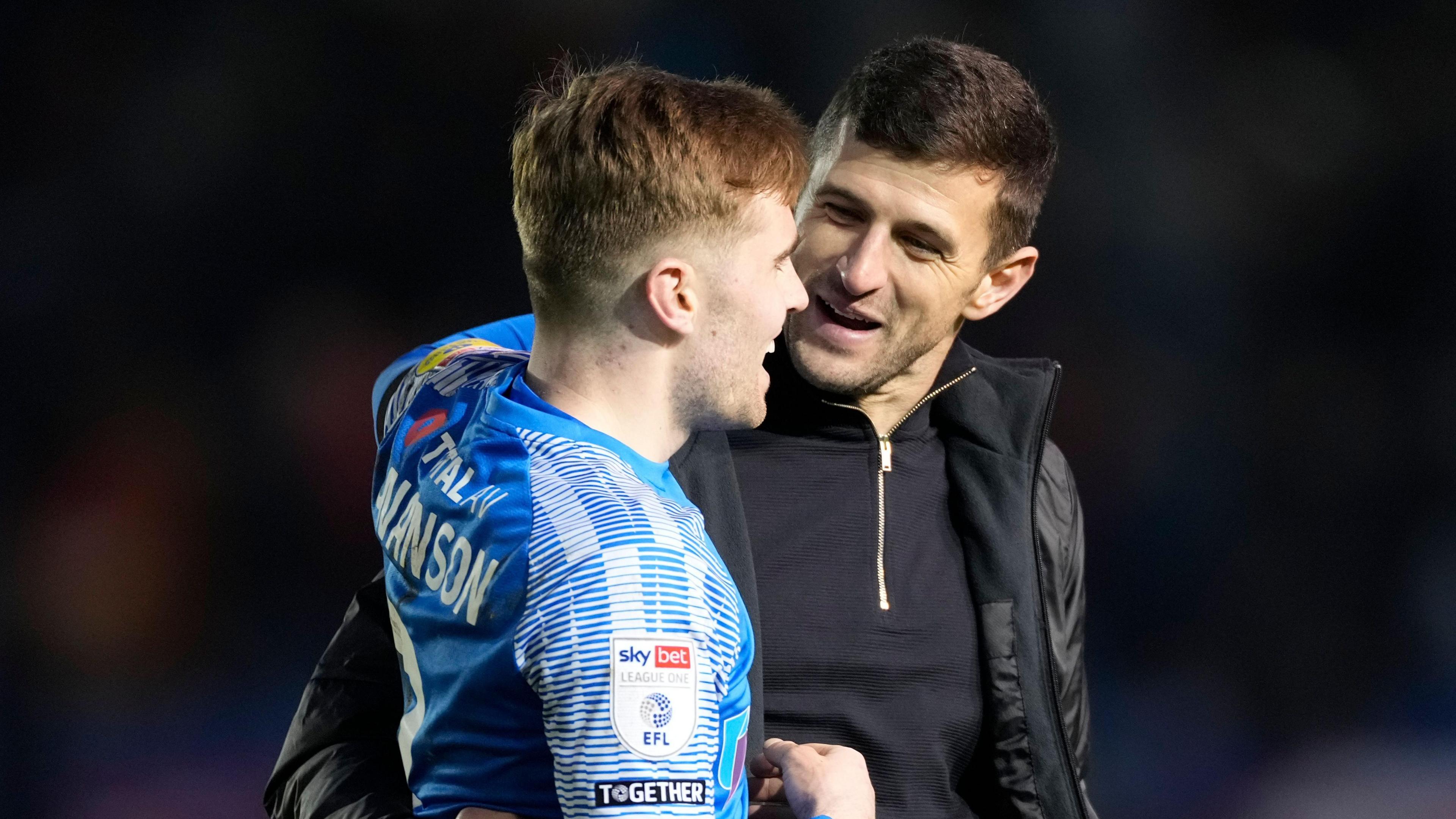 A photo of John Mousinho, wearing a black zip-up jumper and a black coat, embracing Zak Swanson in his blue Portsmouth kit after a match in January 2023