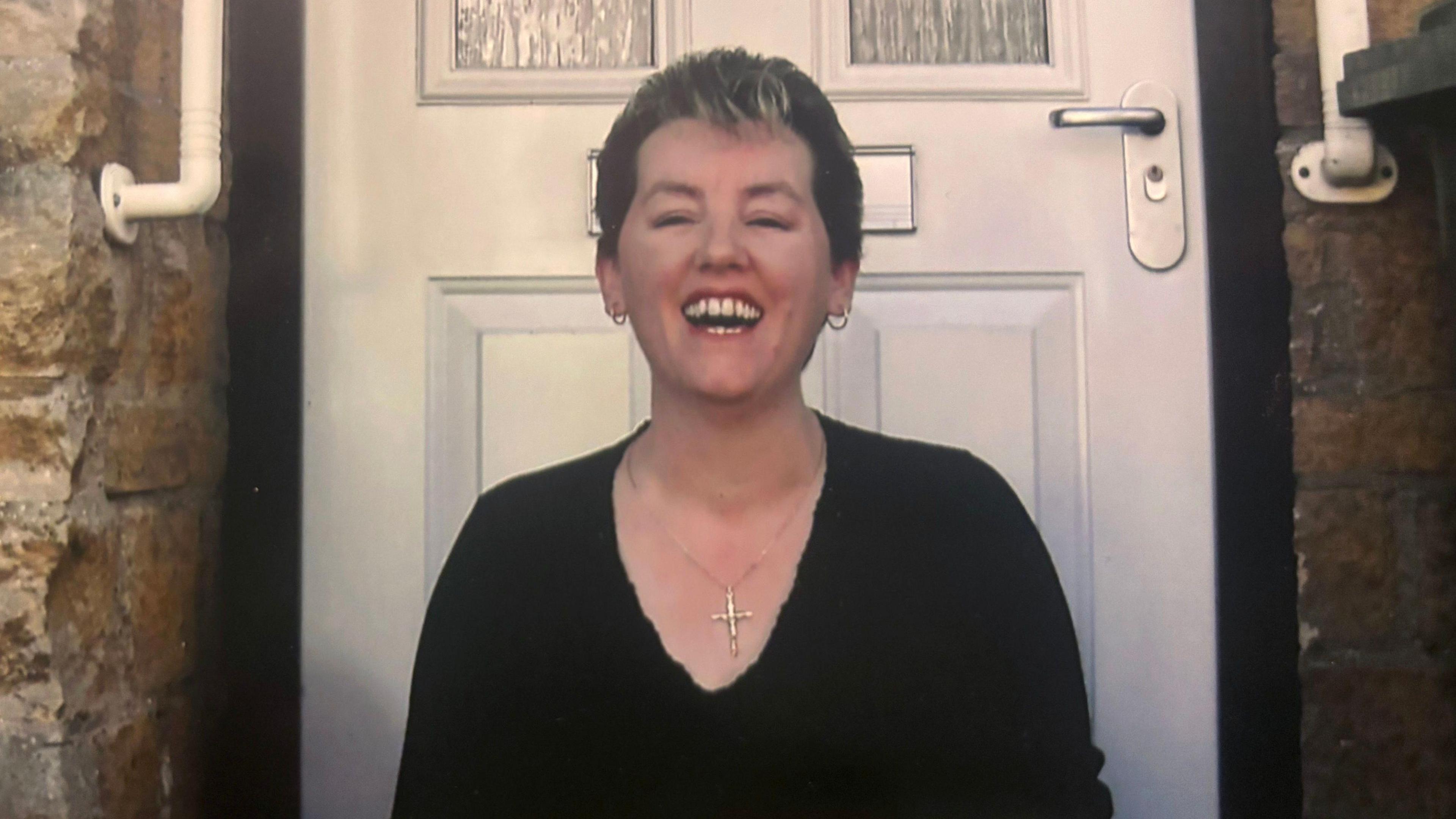 A photo of Lorna Luxton. She is standing in front of a white door. She has short brown hair with blonde highlights and is grinning. She is wearing a black v-neck jumper and a gold cross necklace.