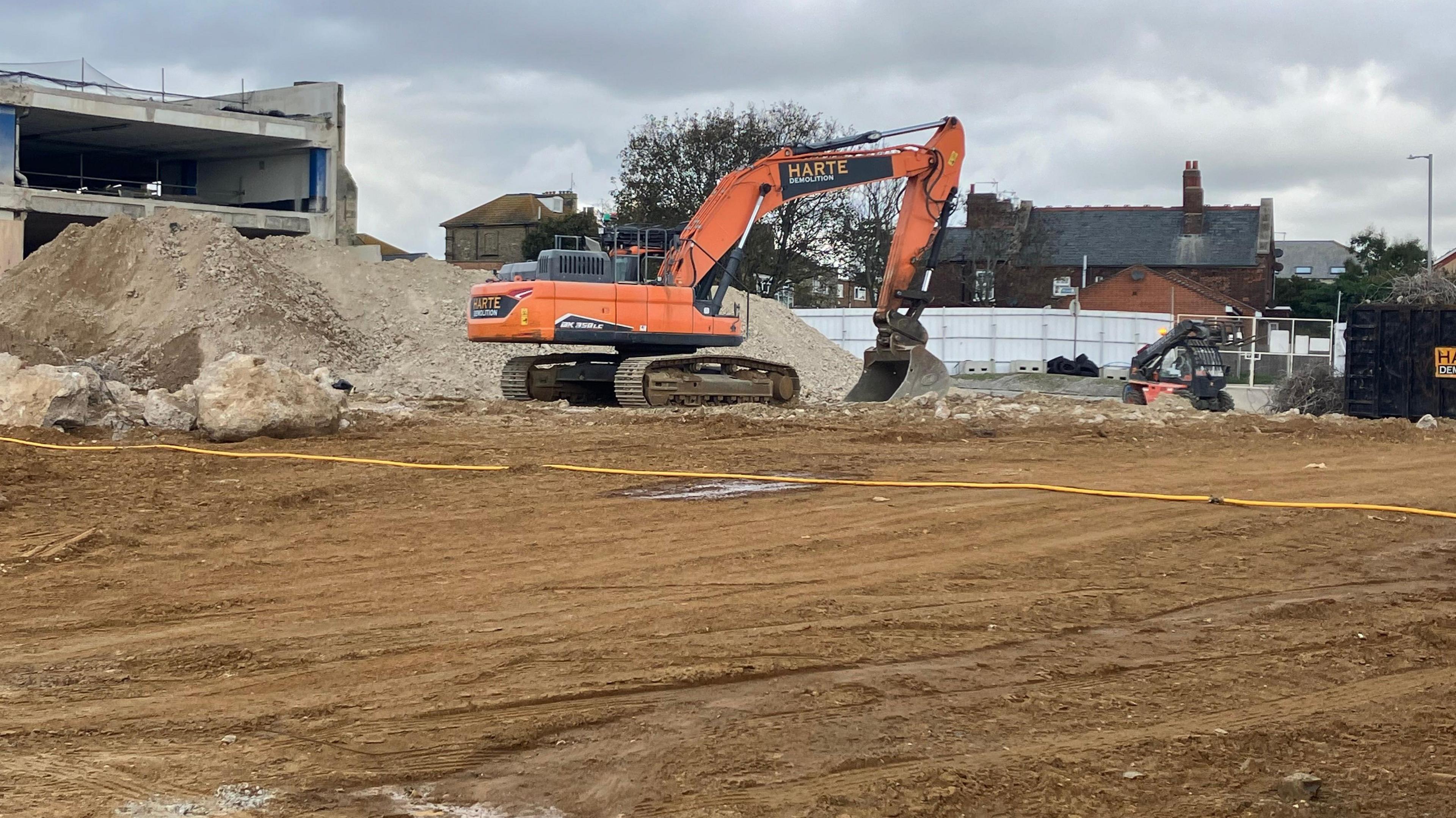 An orange mechanical digger is working on a site of a former car park which is being demolished with a high mound of grey rubble in the background.   