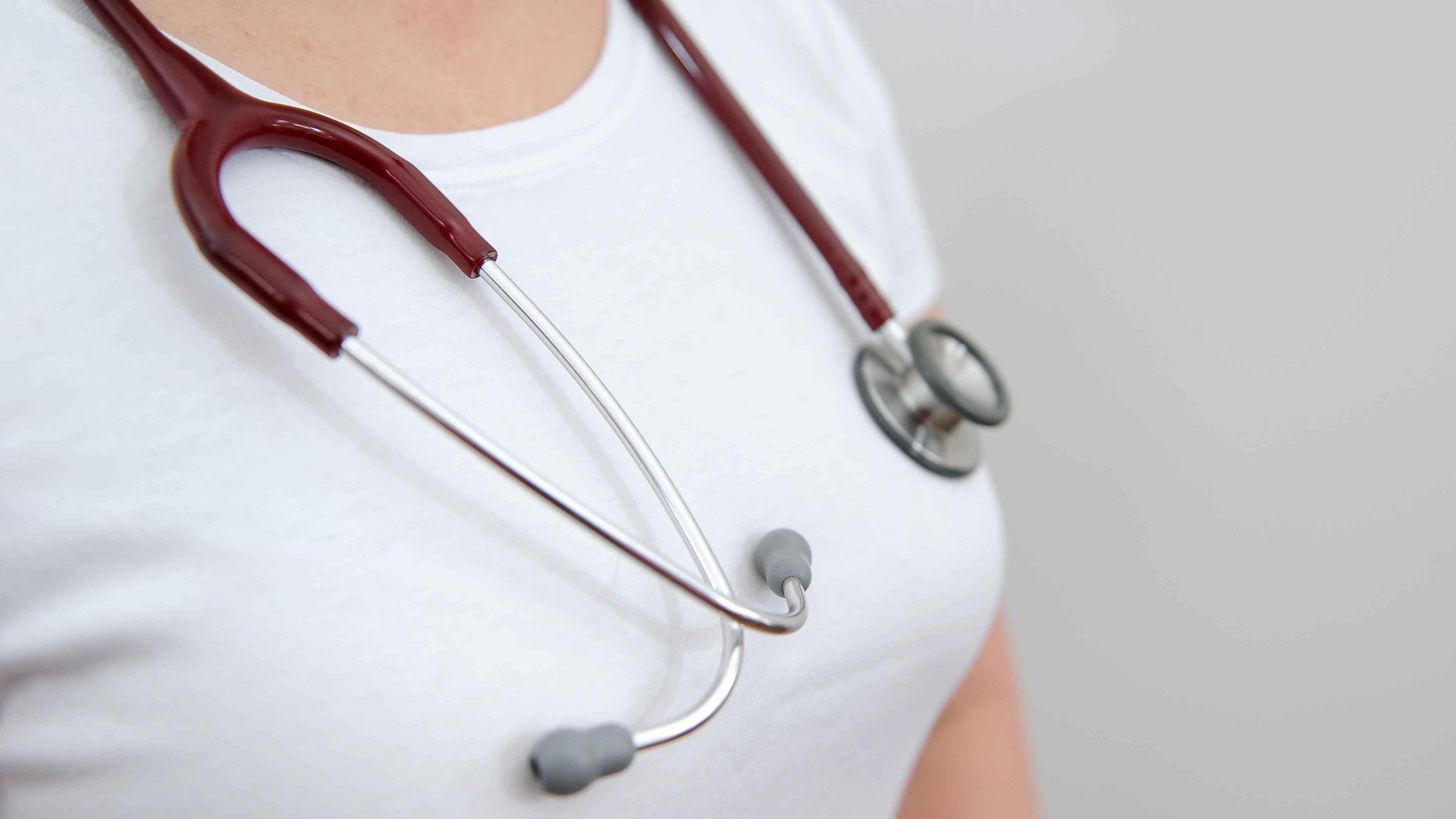 A close-up of a woman wearing a white T-shirt with a stethoscope around her neck.