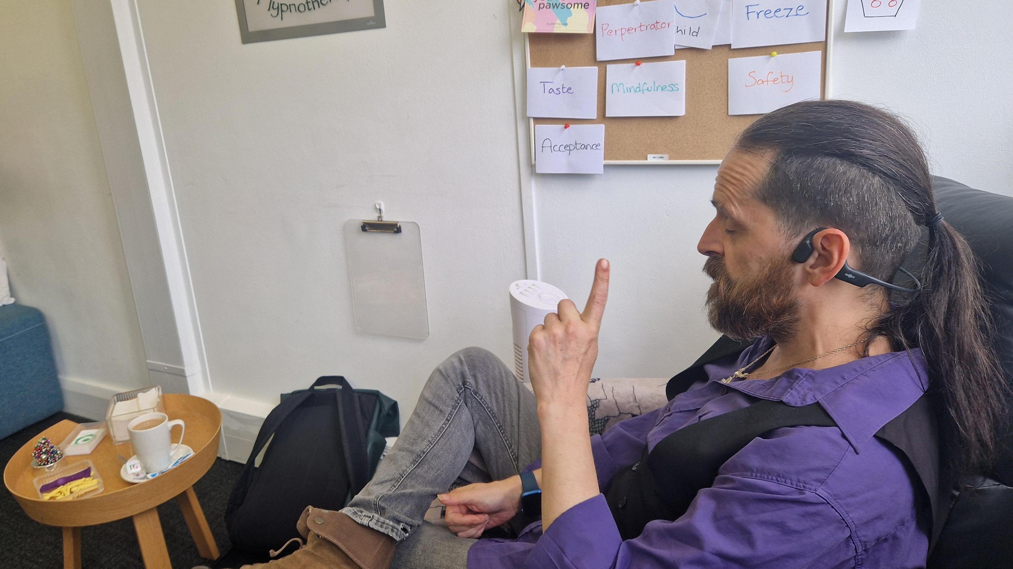 Rob has dark hair tied back in a pony tail and a beard. He is wearing a purple shirt which is open at the collar, a black waistcoat and a large silver pendant necklace. He is sitting in his office and chatting. He is to the right of the photo, above him is a notice board with various white notes on it. There is a coffee table in front with a white mug, which has a cup of tea in it. 