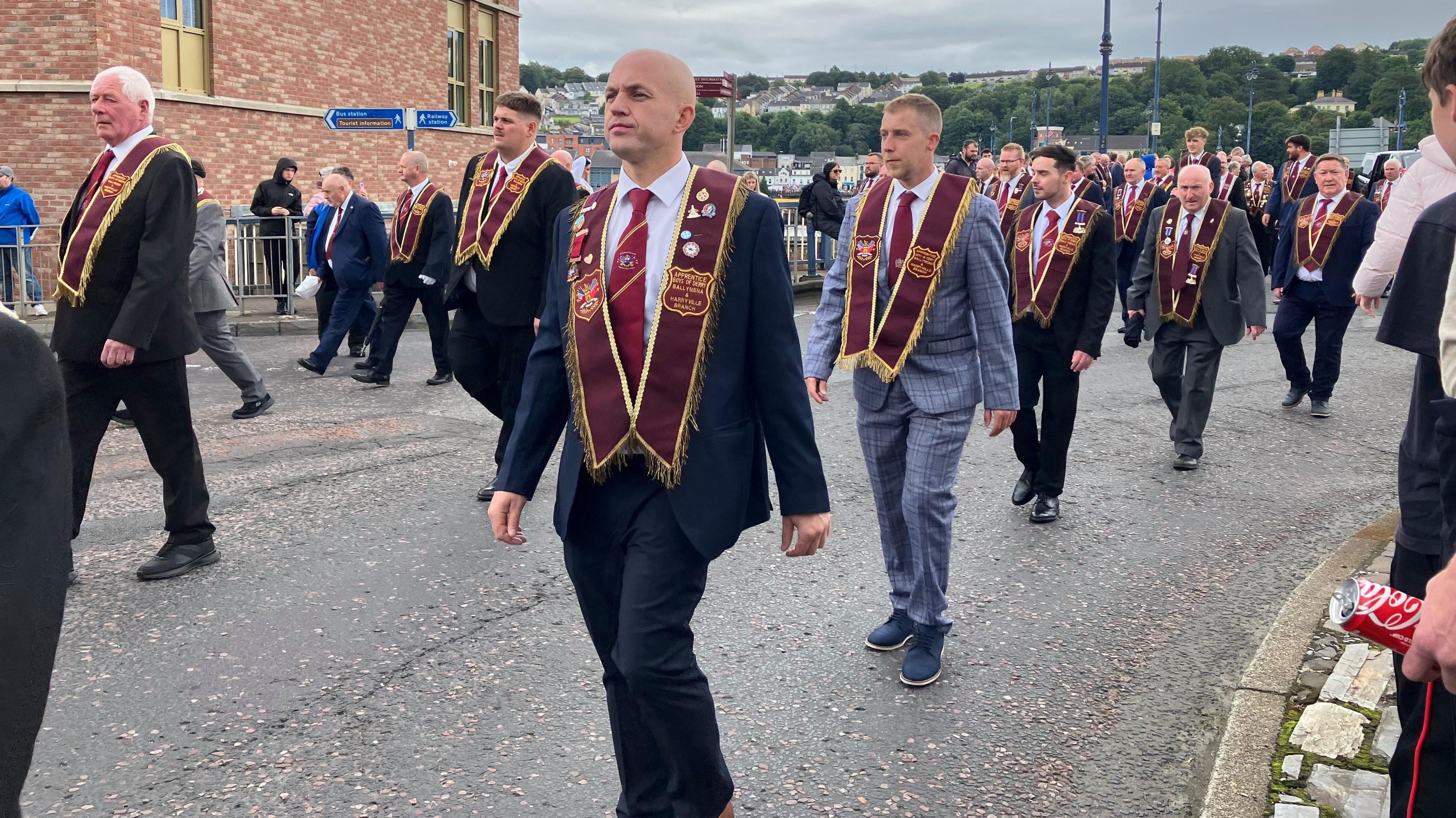A group of men wearing black and grey suits marching in a parade. They are wearing maroon sashes an carrying various flags.