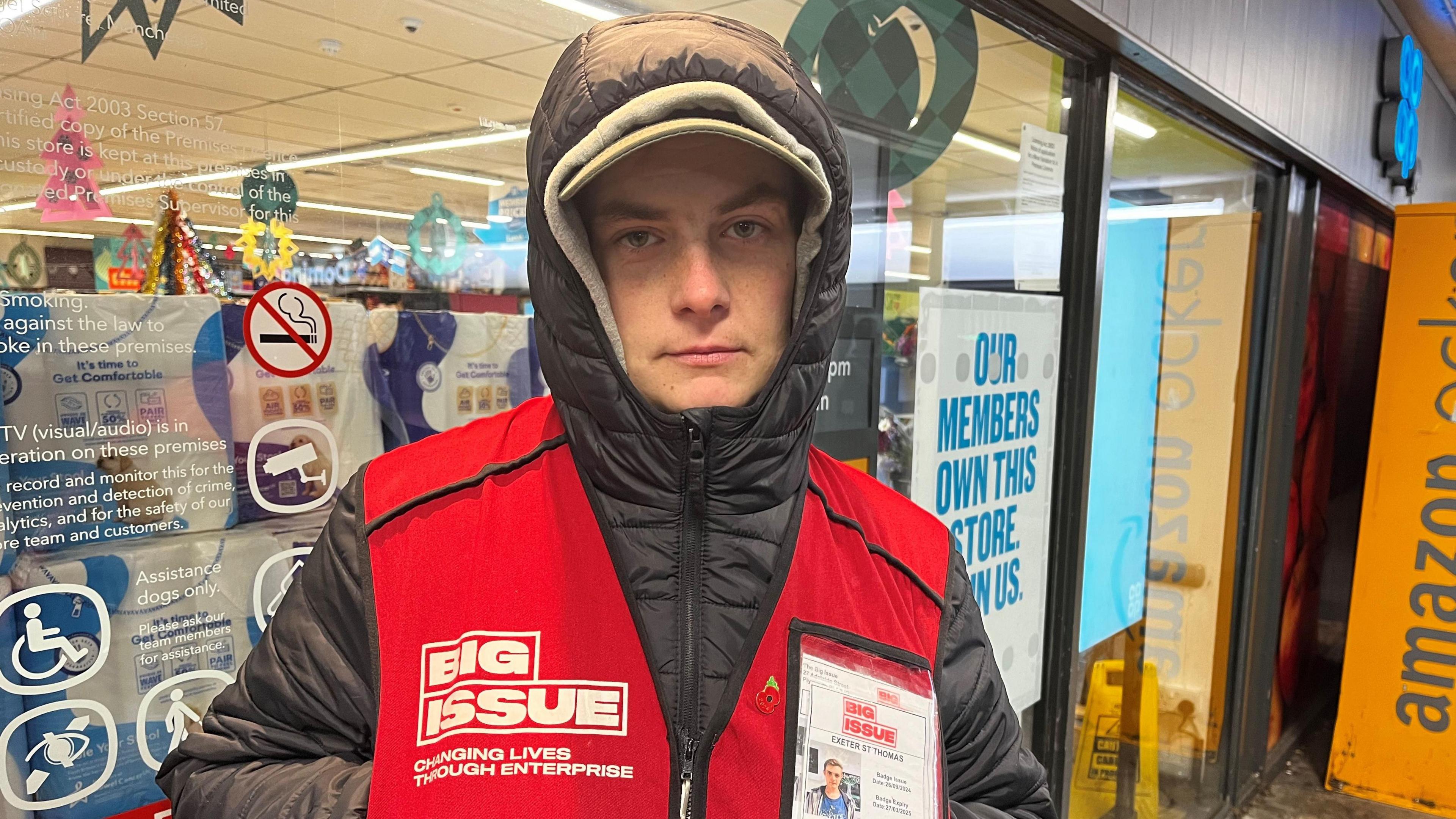 Former rough sleeper Alfie stands outside a Co-op store in Exeter. He is wearing a black puffer jacket with the zip fully done up and the hood up over his head. He also has a red Big Issue jacket on. Christmas decorations can be seen inside the store behind Alfie. He is stood near a yellow Amazon delivery storage unit.