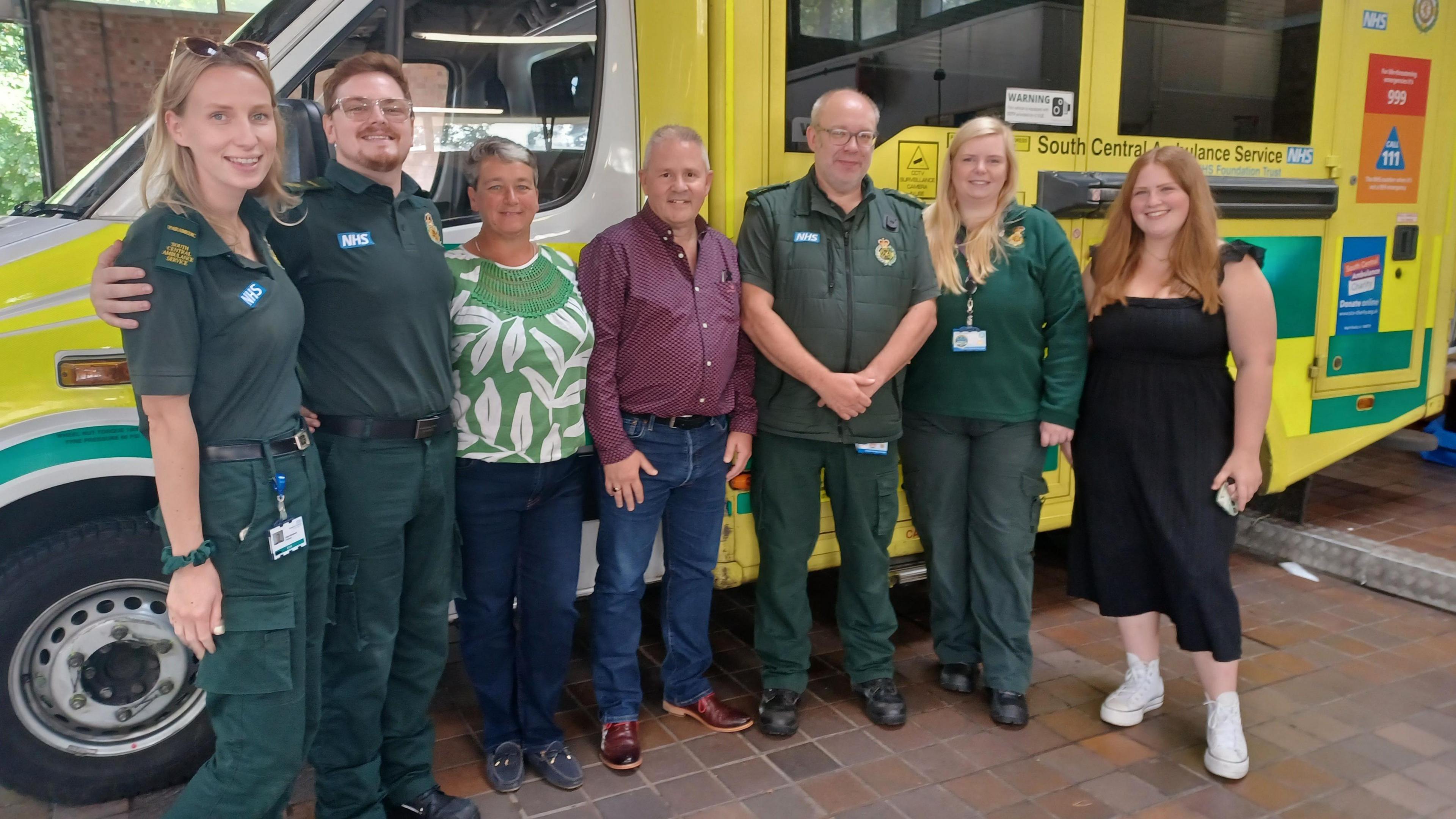 Barry Crawford, a man in blue jeans and a red shirt, and his wife Alison, wearing a green top and jeans, stand with four paramedics in front of an ambulance.