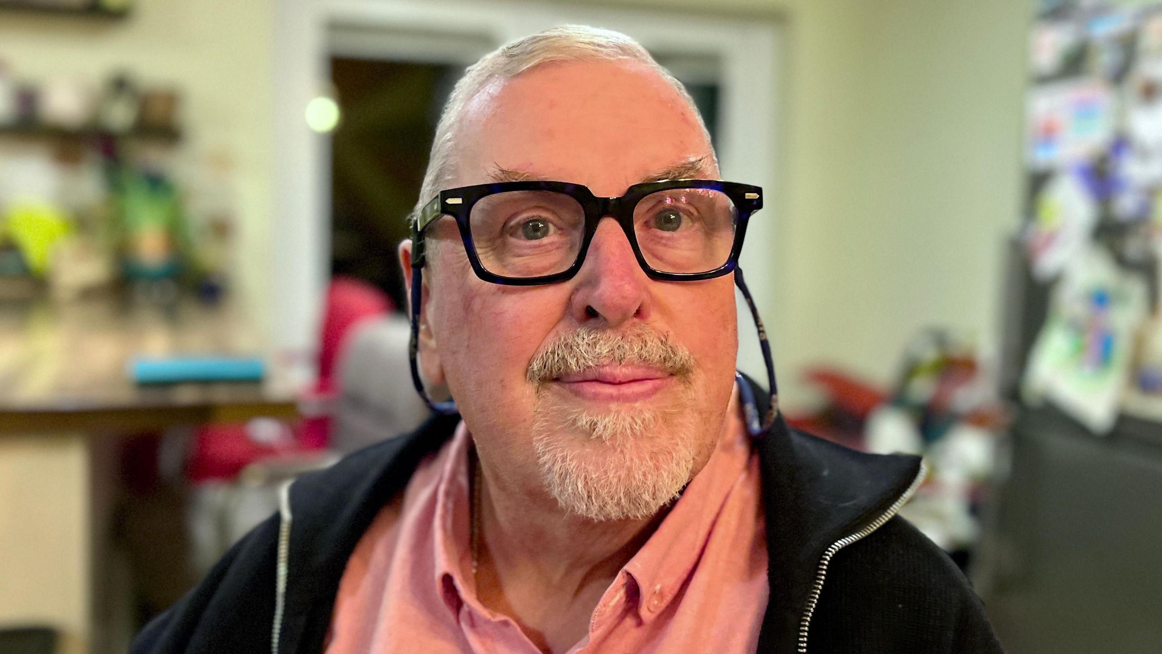 Steve O'Farrell looks directly into a camera. You can see his head and shoulders. He is wearing glasses, a pink shirt and black jacket. The background is blurred, but he appears to be sat in a kitchen.