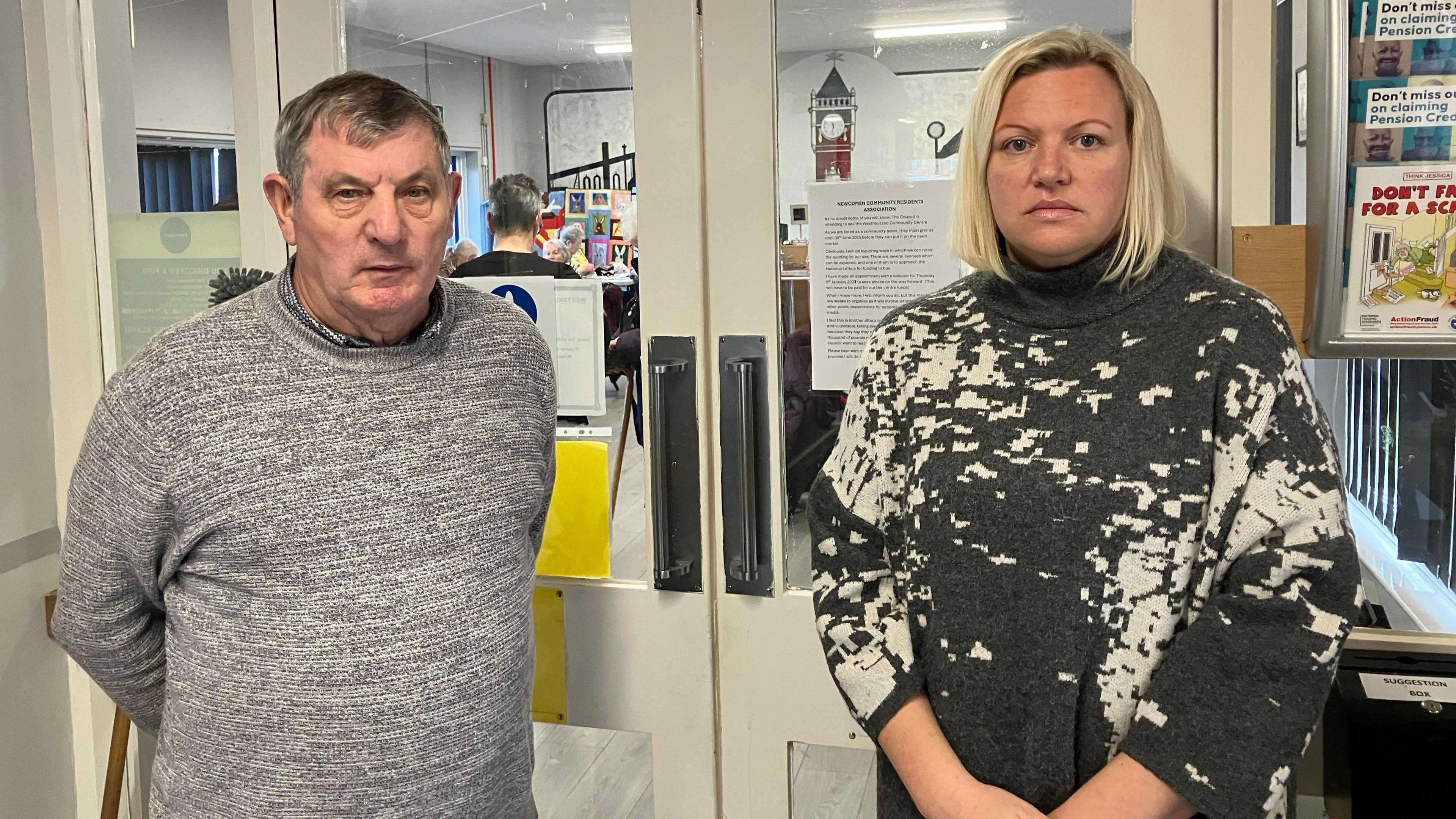 Trustee Billy Wells and councillor Carrie Richardson stand in front of doors to the community centre. He has short grey hair and wears a grey jumper. She has blonde hair in a bob cut and wears a grey and white jumper.