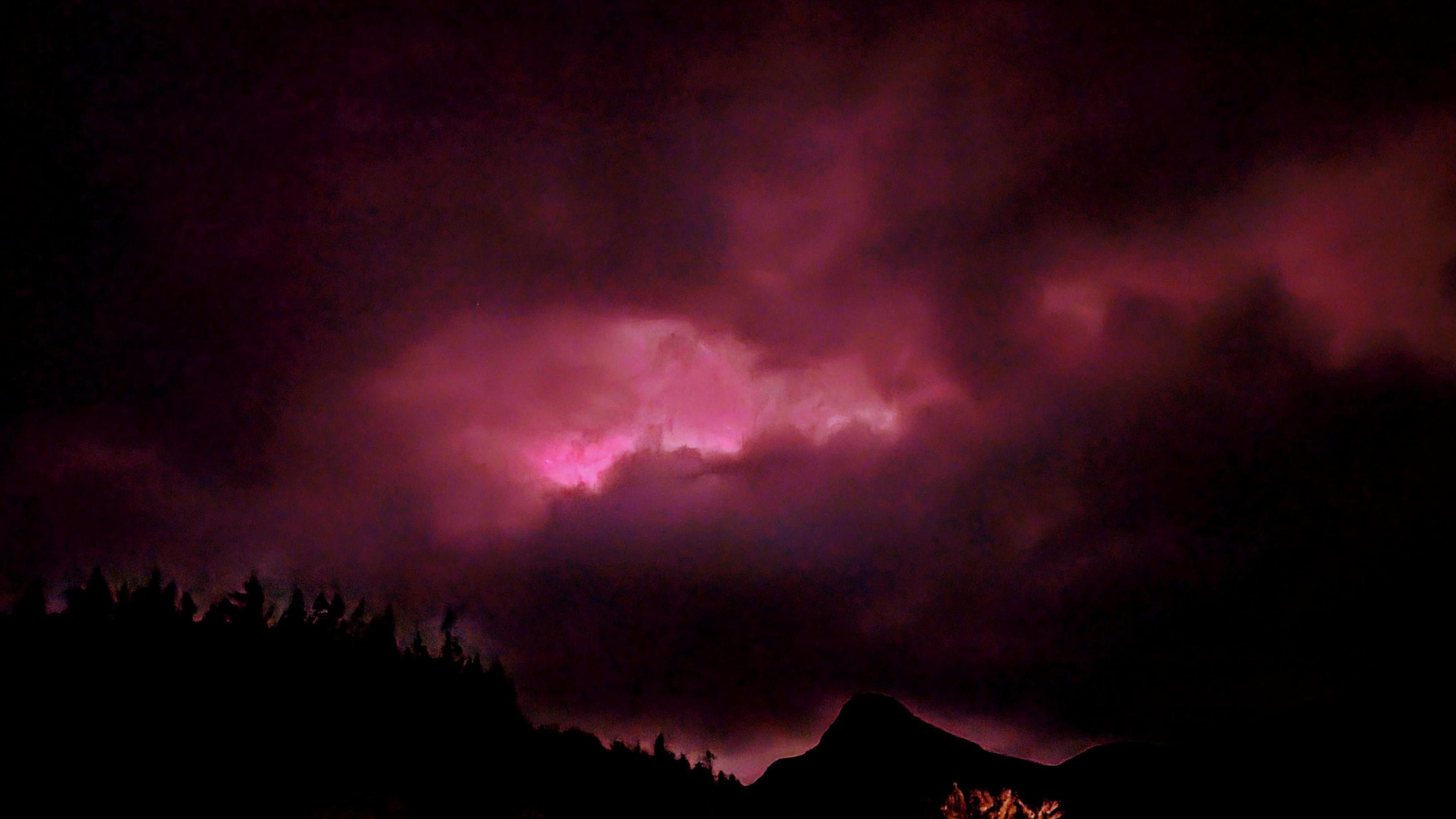 Aurora appears pink in colour behind clouds and a hill.