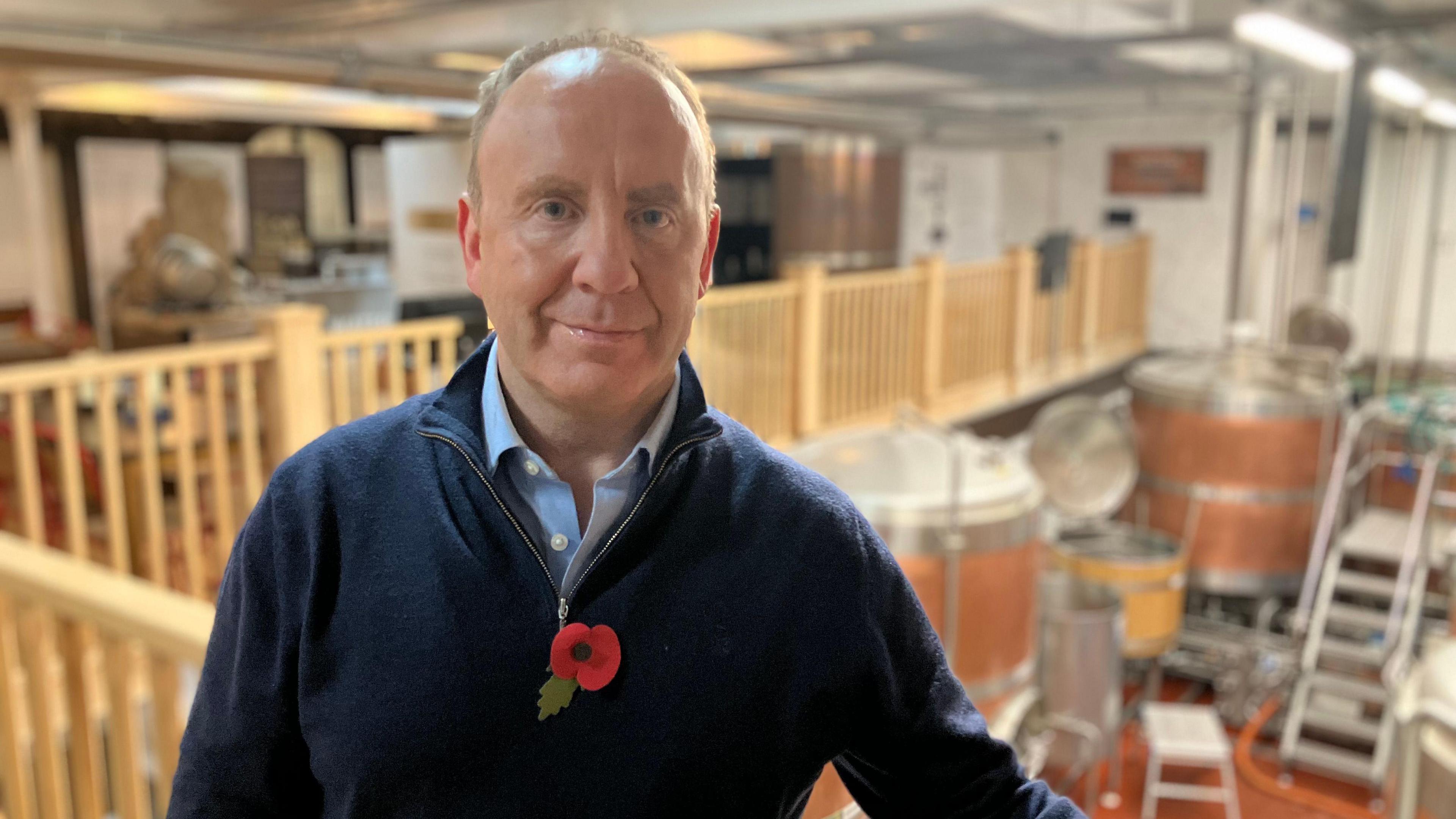 A man stands in a brewery in front of large vats that are used to brew beer. He is wearing a blue jumper and a red Remembrance Day poppy. 