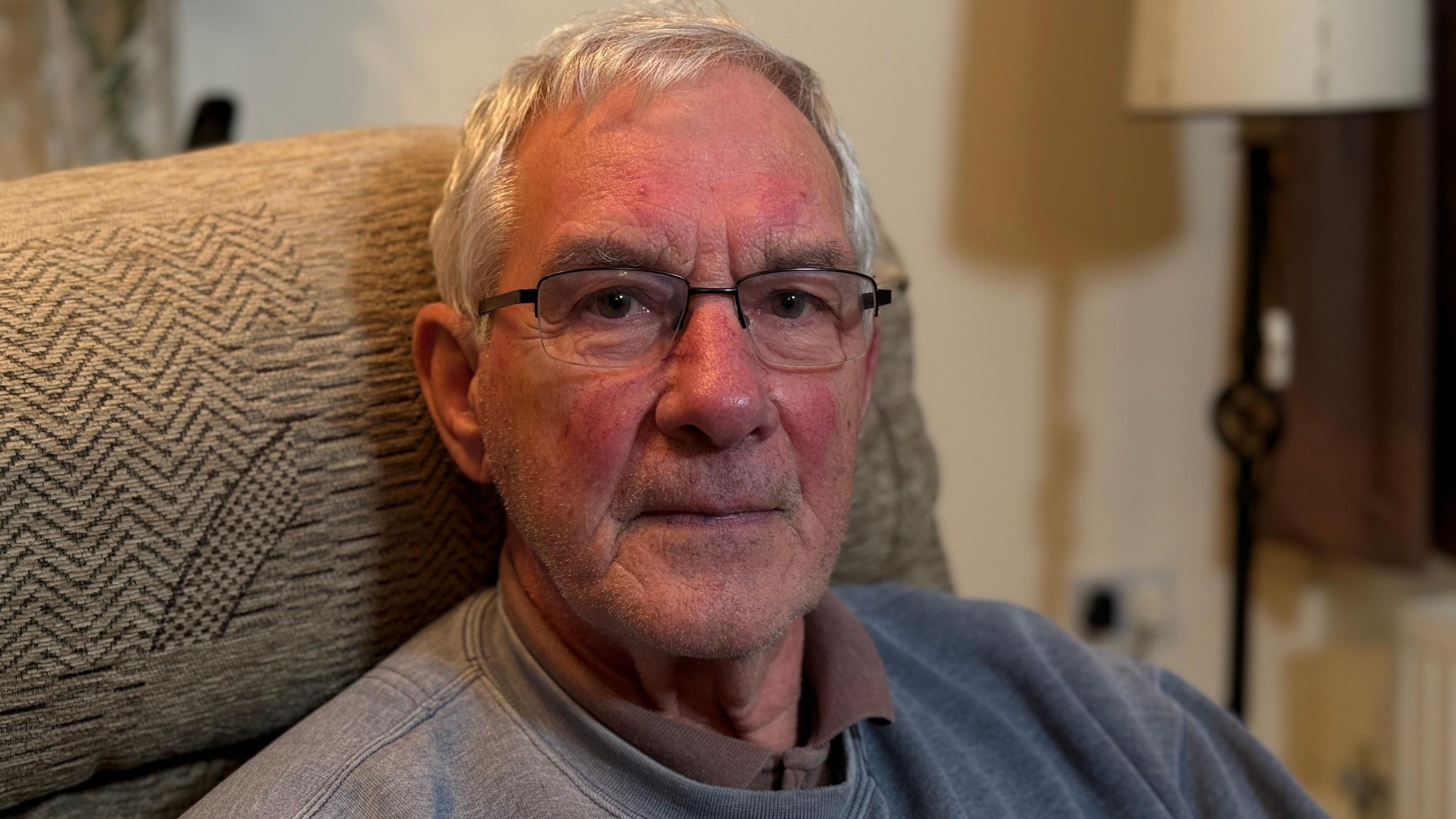 John Bolton, who has grey hair and black-framed glasses and is wearing a grey jumper, sat on a beige sofa looking left of the camera