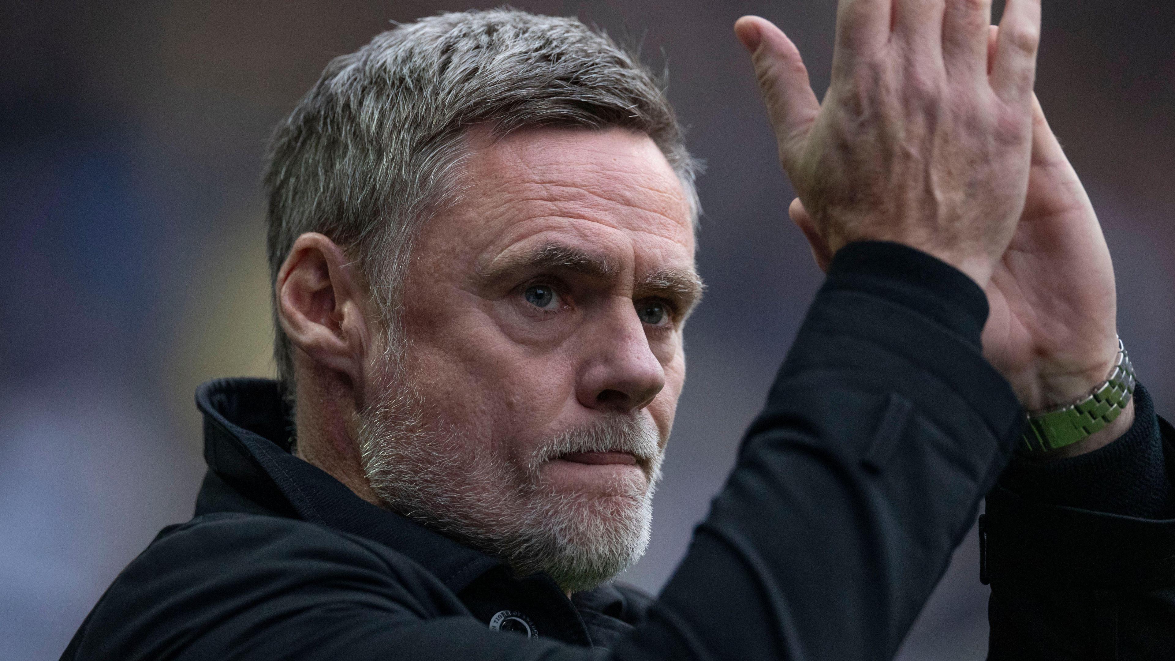 A close up of Graham Alexander applauding wearing a long-sleeved black jacket