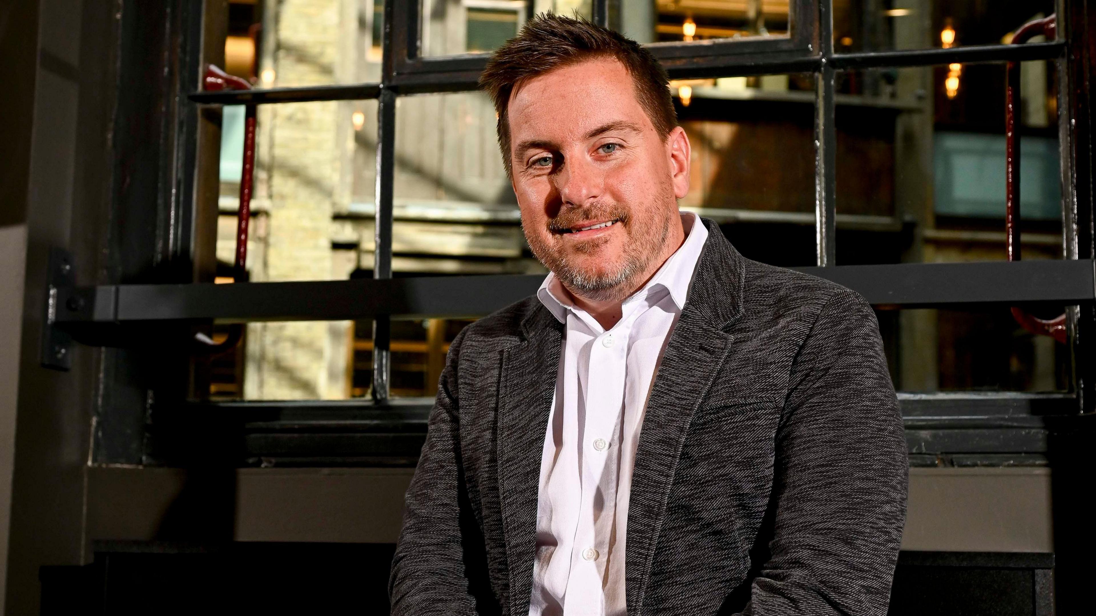 Peter Taylor sitting down in a room, with a window behind him. He is looking at the camera, smiling, wearing a pink shirt, dark jacket, he has short dark hair and a stubbly beard. 