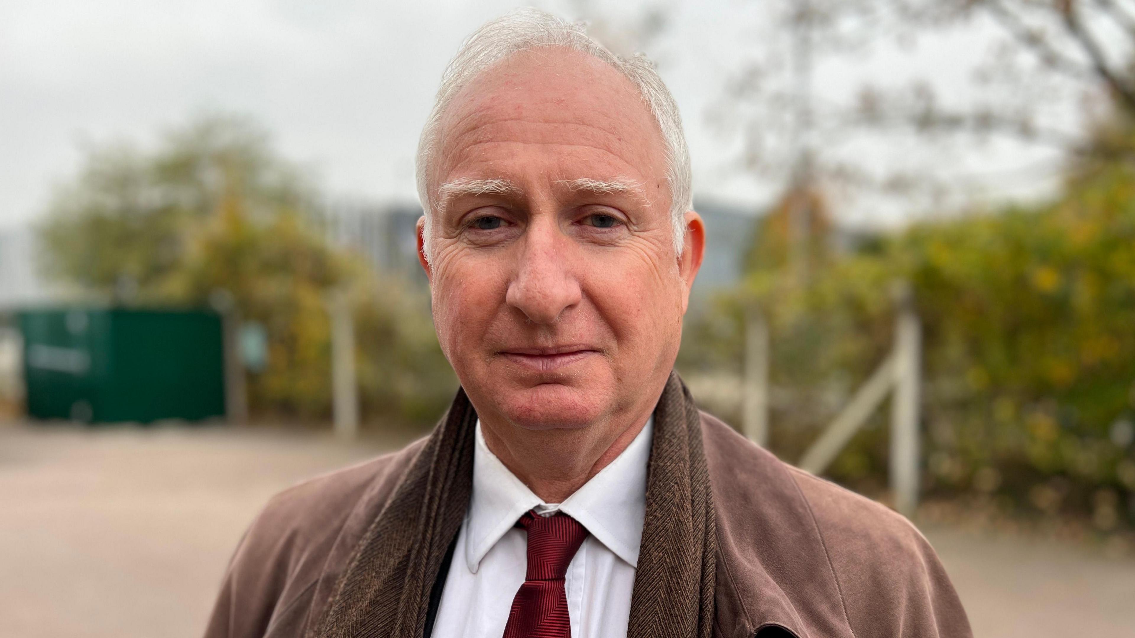 Daniel Zeichner, a man wearing a white shirt, red tie and brown jacket. He is standing outside and is looking into the camera.