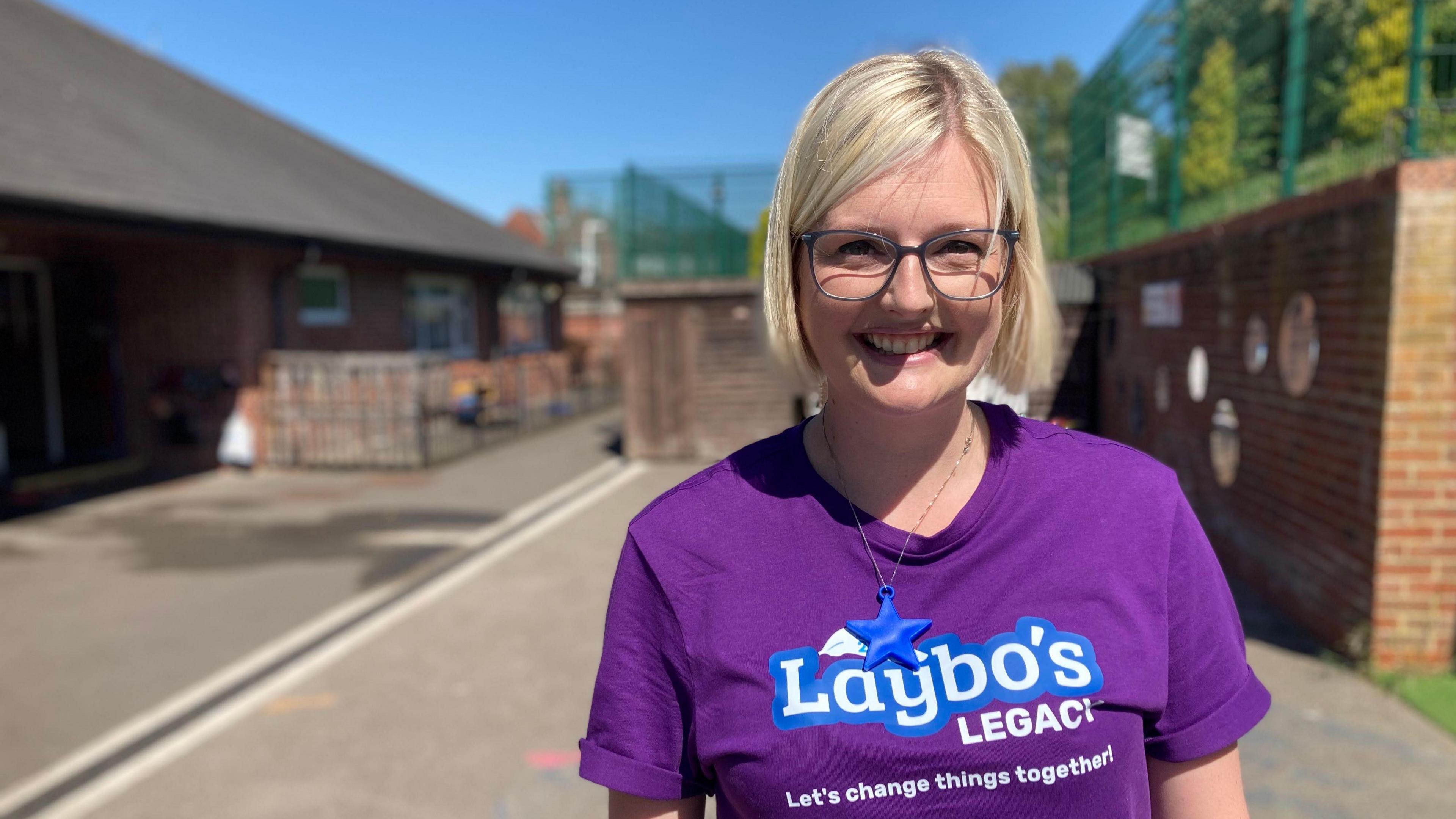 Emily Kitchener. She has short blond hair and is wearing glasses and a purple T-shirt with the words Laybo's Legacy - let's change things together.