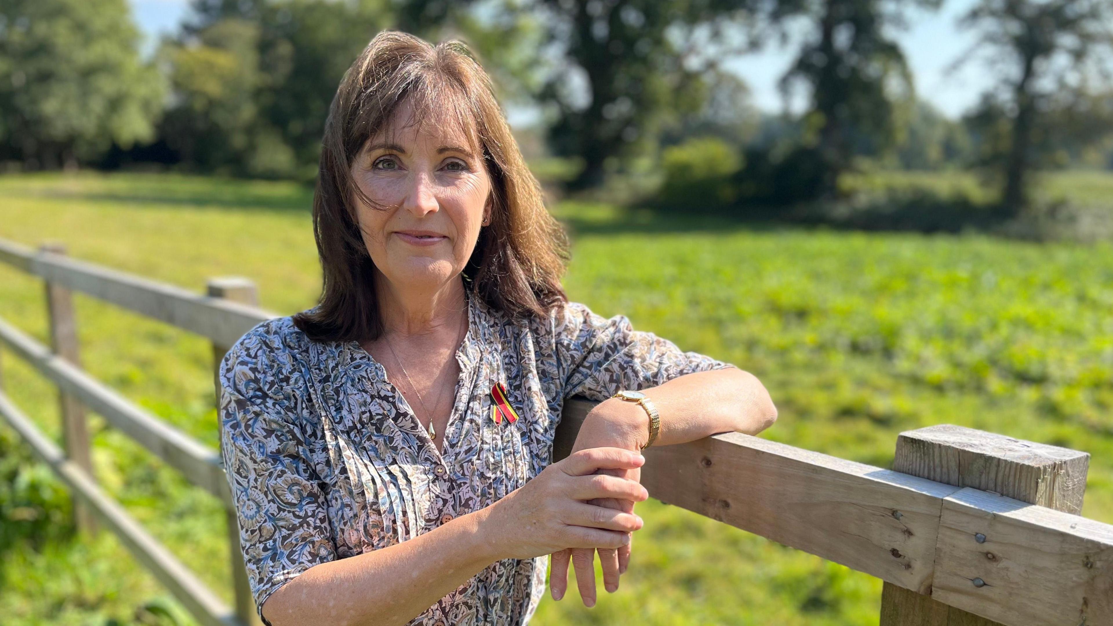 Amanda Cummings leans her left arm on top of a wooden fence with a green field and trees in the background. She has shoulder length brown hair with a fringe and is wearing a light coloured patterned blouse with a red ribbon pinned to the top left. She is looking into the camera with a small closed mouth smile