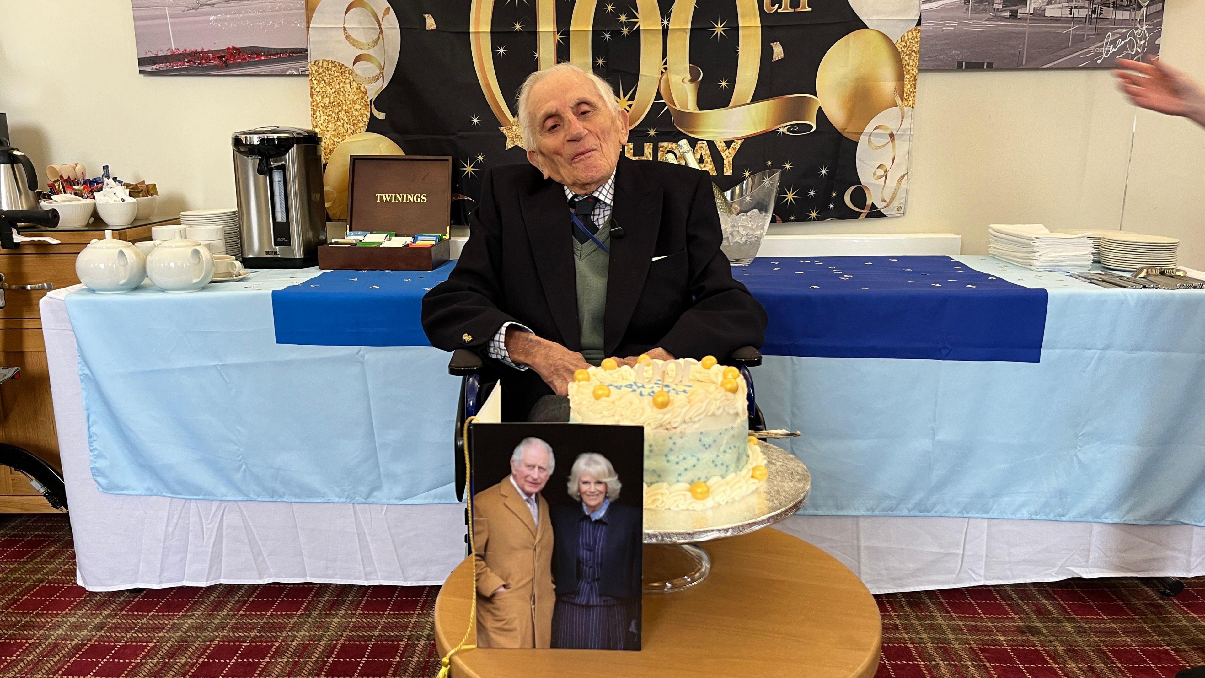 Captain Walter Godsal is sitting in front of a 100th Birthday banner. A tiered cake is on a table in front of him.