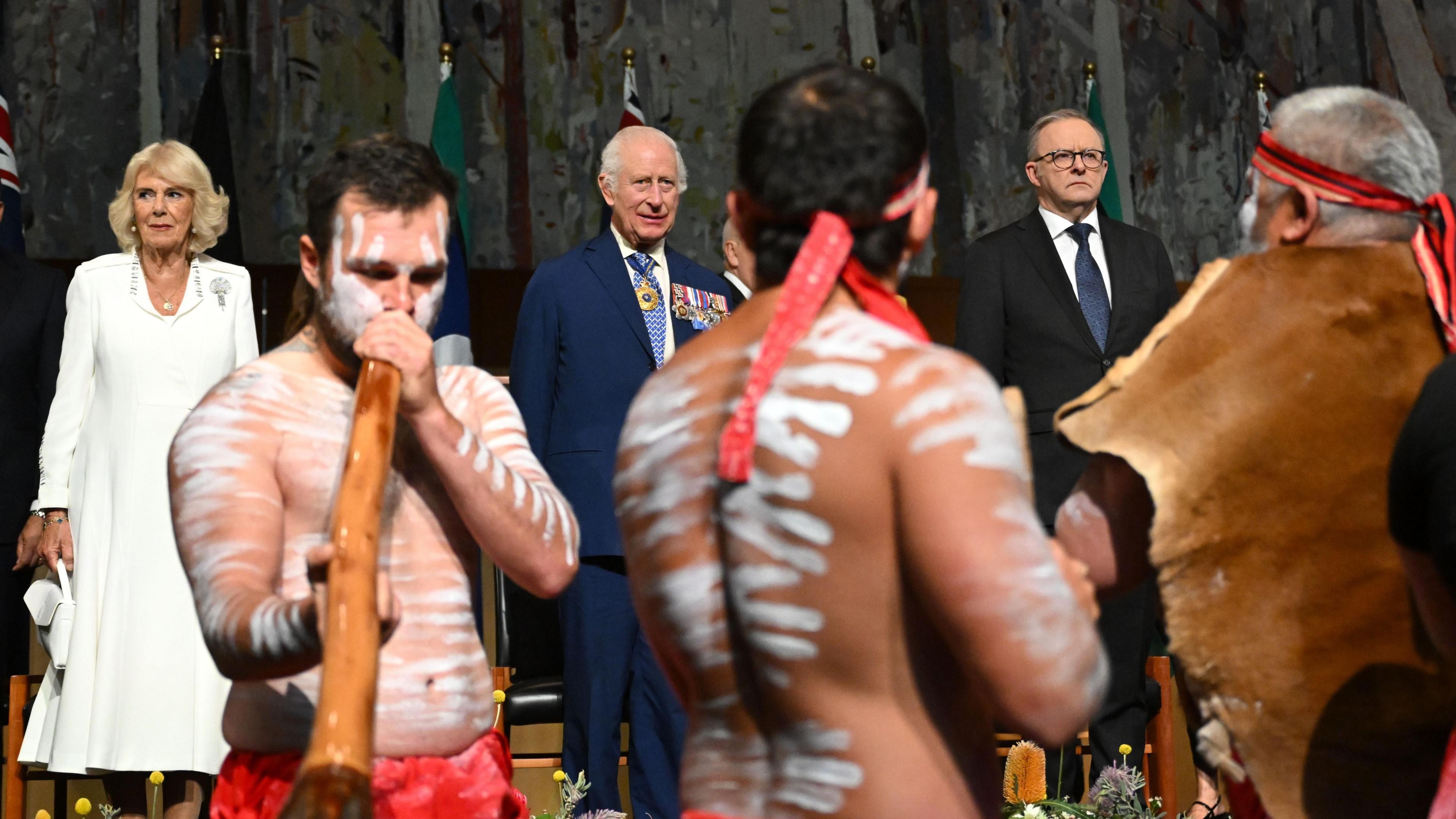 Three men in traditional Aboriginal clothing and body paint play digeridoos watched on by Queen Camilla, King Charles and Australian Prime Minister Anthony Albanese