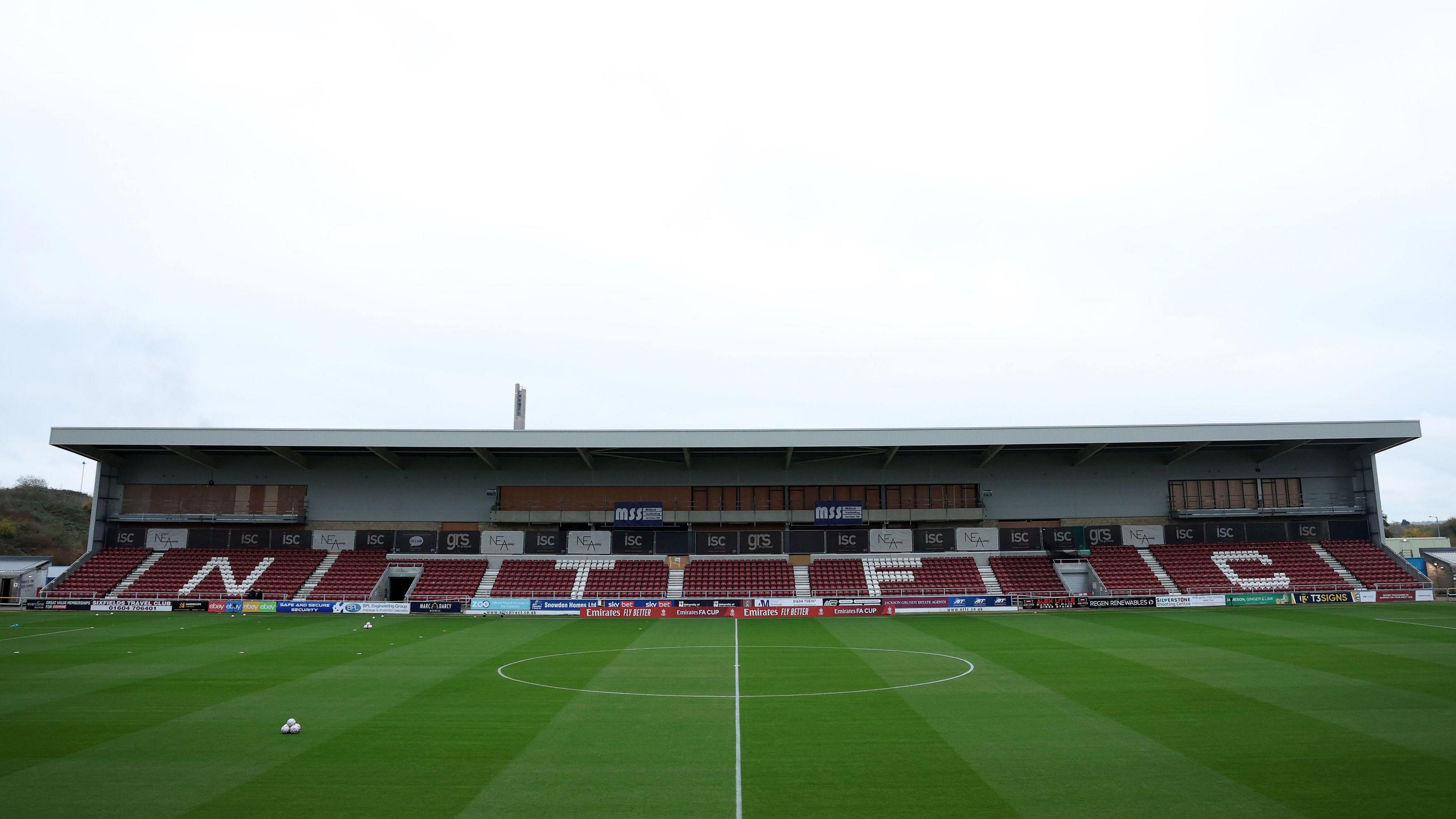 A wide shot if the Northampton Town football pitch and stadium. The stadium is currently empty with claret and white seats. 