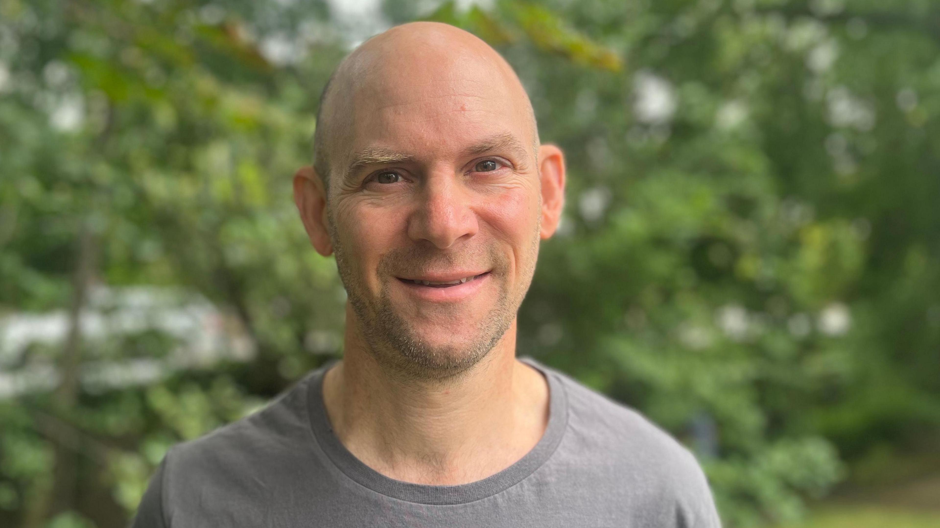 A man in a grey T-shirt stands in front of several trees and hedges. He is looking directly at the camera and smiling.