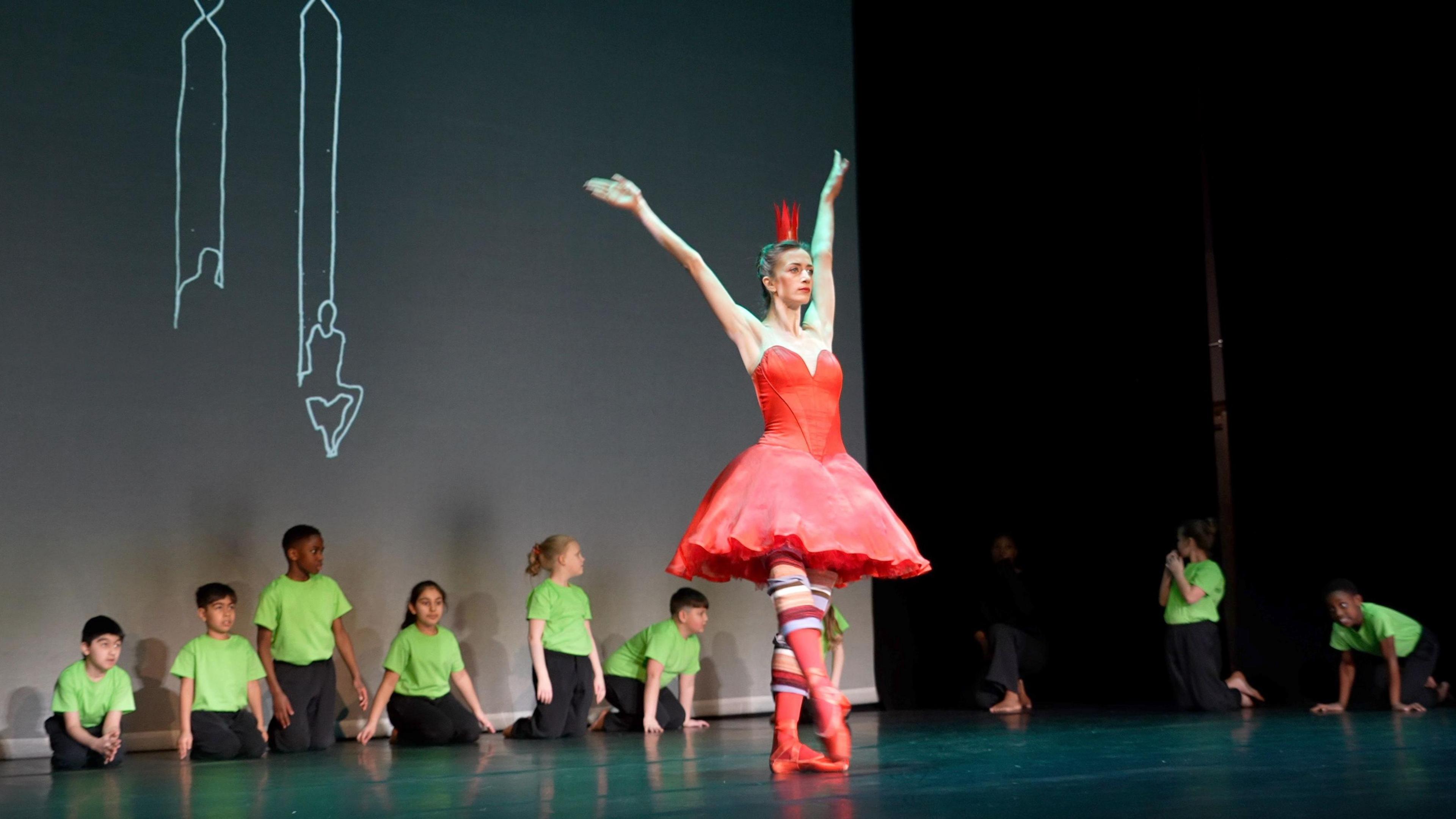 A woman in a red costume stands on a black stage with children behind her wearing black trousers and green T-shirts.