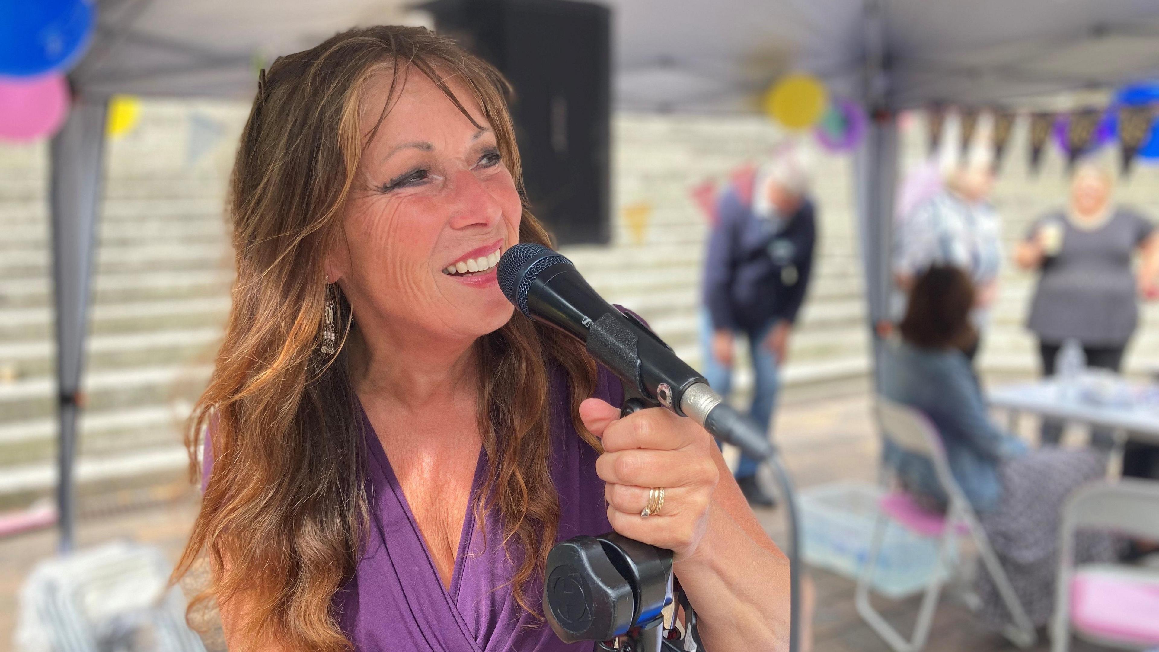 A lady with long red hair and wearing a purple dress singing into a microphone with a marquee, bunting and some people in soft focus behind her.