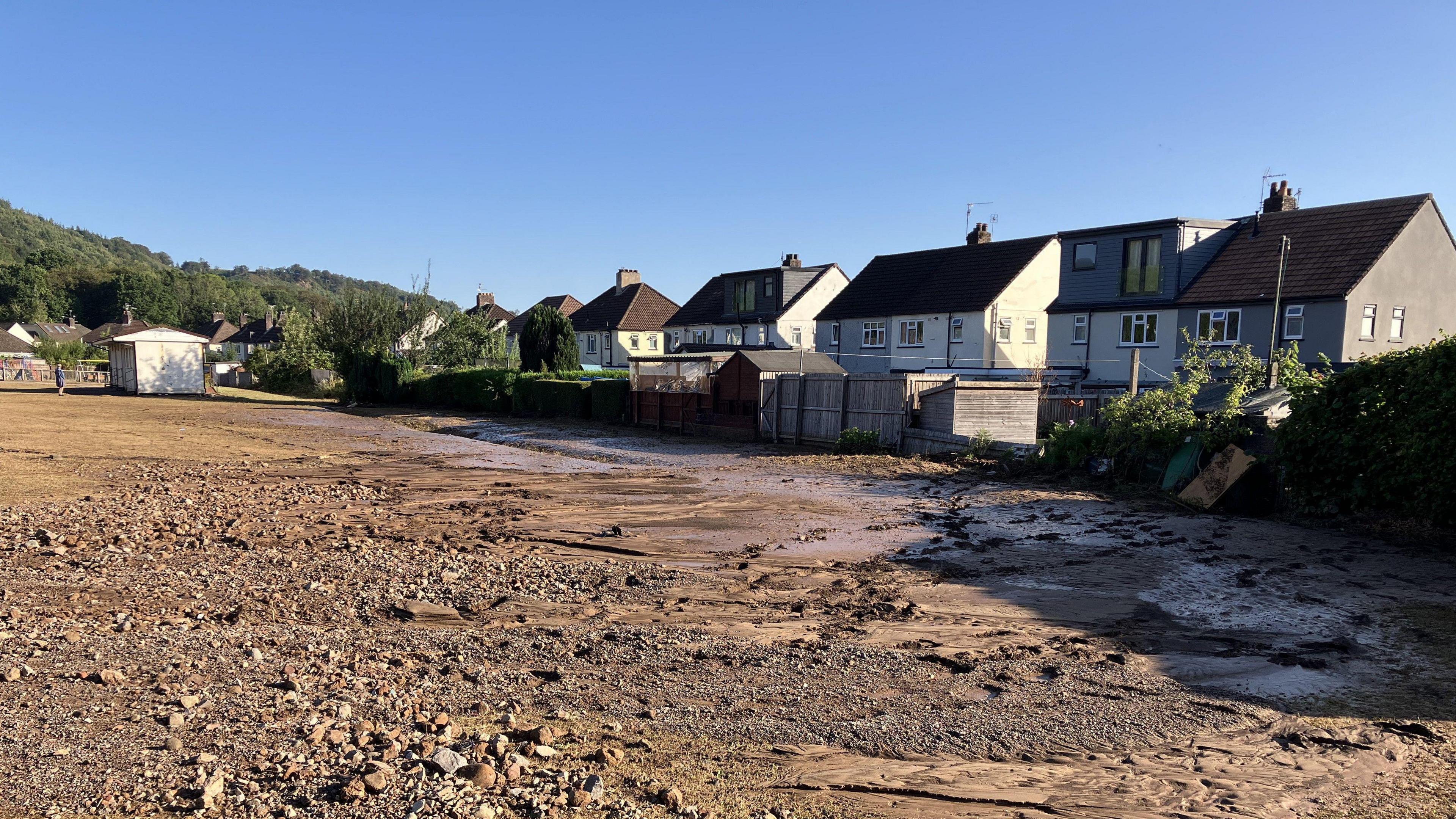 A football pitch which is covered in wet mud and stone sits with houses on Heol Berry backing on to it. 
