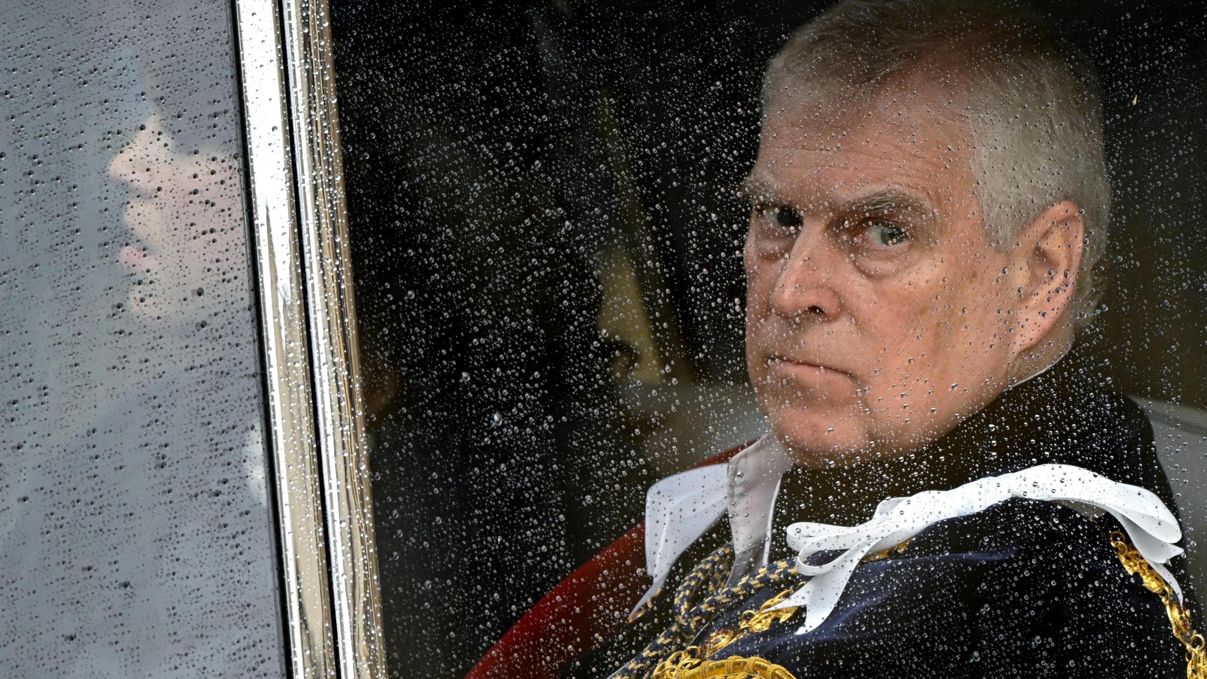 Prince Andrew looks through a carriage window, wearing a black jacket. There is rain on the window.