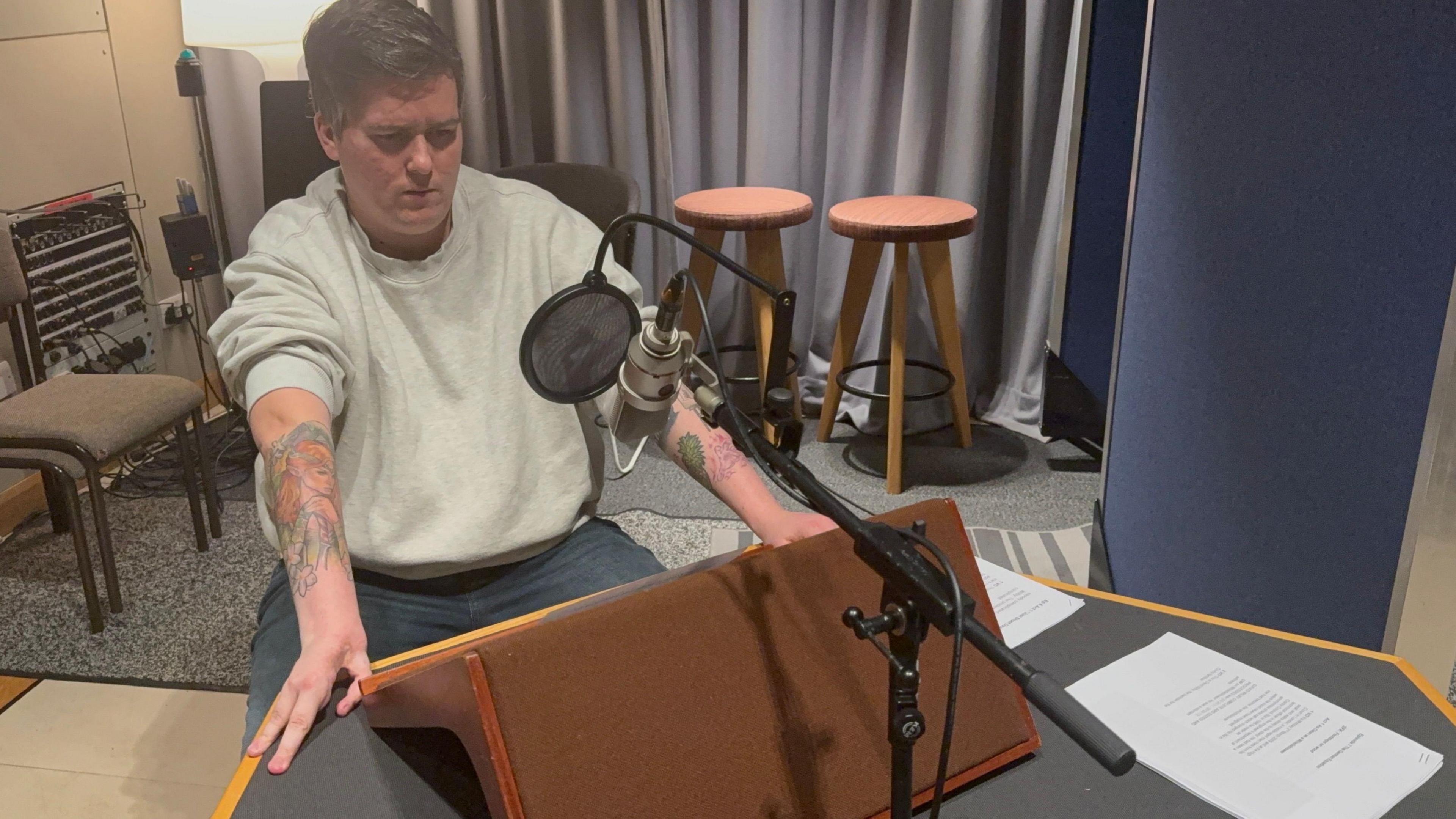 A man in a grey sweatshirt with the sleeves rolled up sits in a recording booth, reading lines into a microphone.  