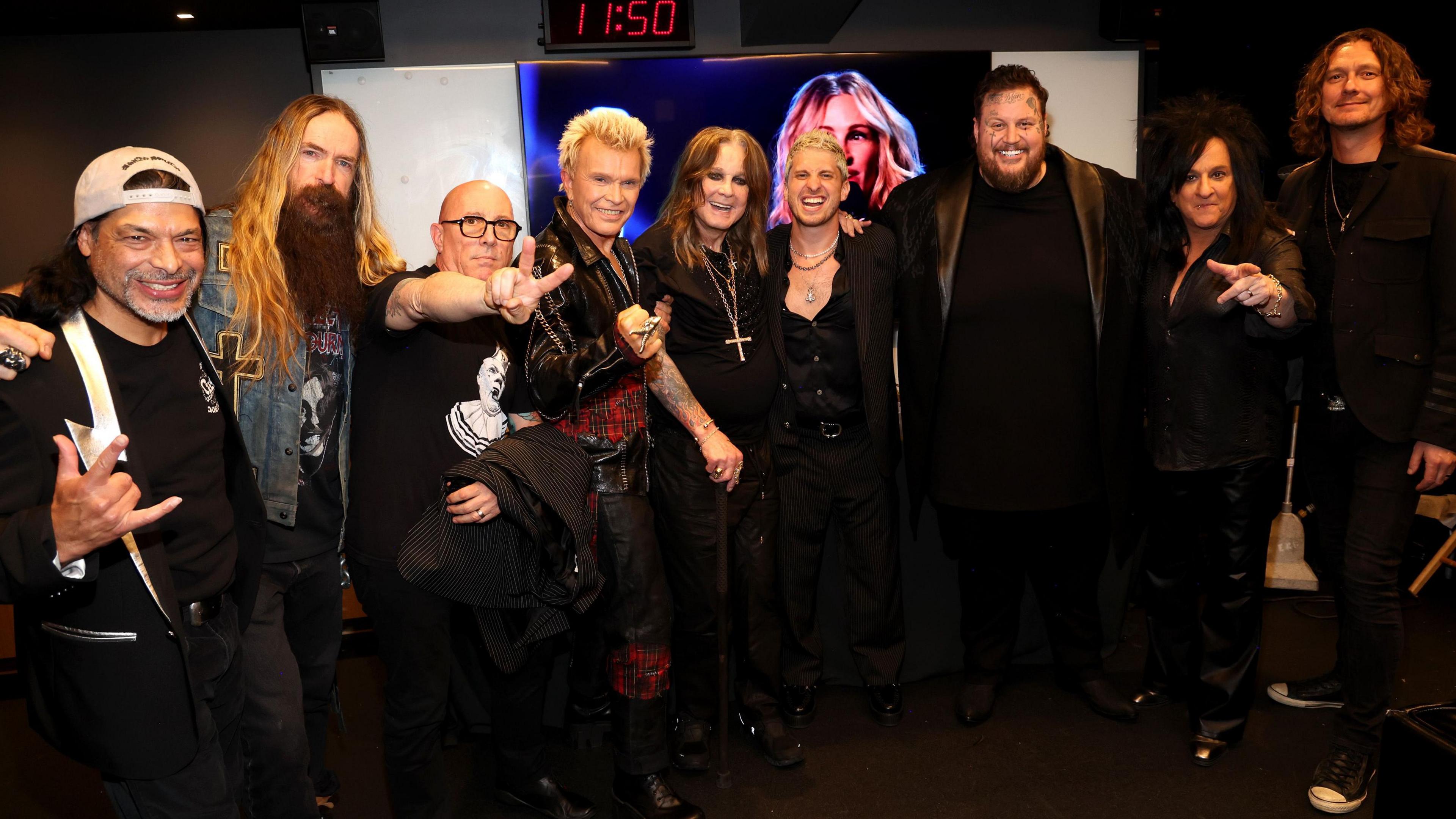 A group of nine men smiling at the camera. They are wearing mainly black outfits.