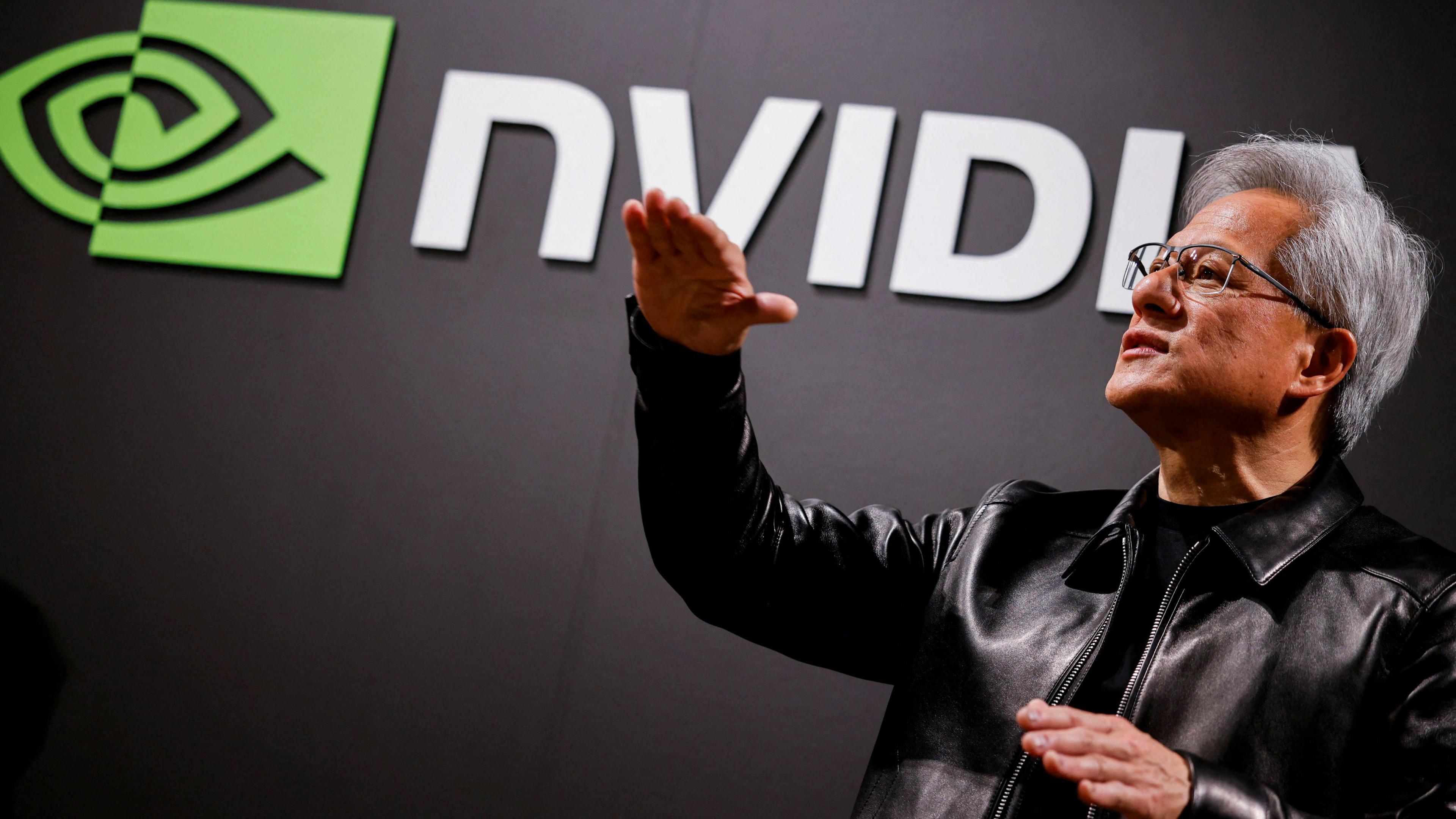 Nvidia CEO Jensen Huang standing in front of a screen with the company's name and logo, gesturing upward with his hands, wearing a black leather jacket over a black shirt.