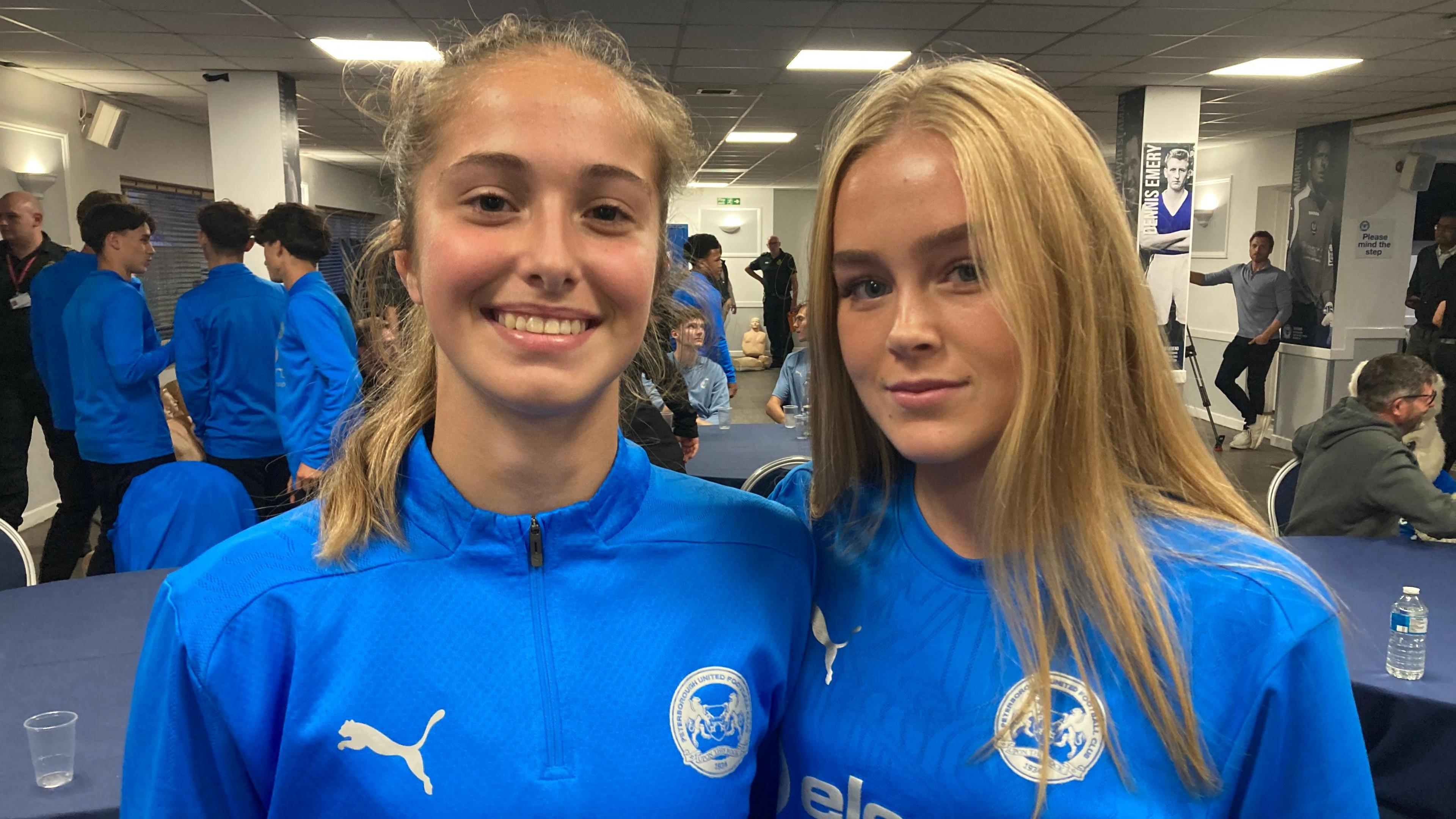 Jess Abrantes and Katie Gee smiling at the camera wearing blue Peterborough United strips. Other players in blue strips can be seen behind them.