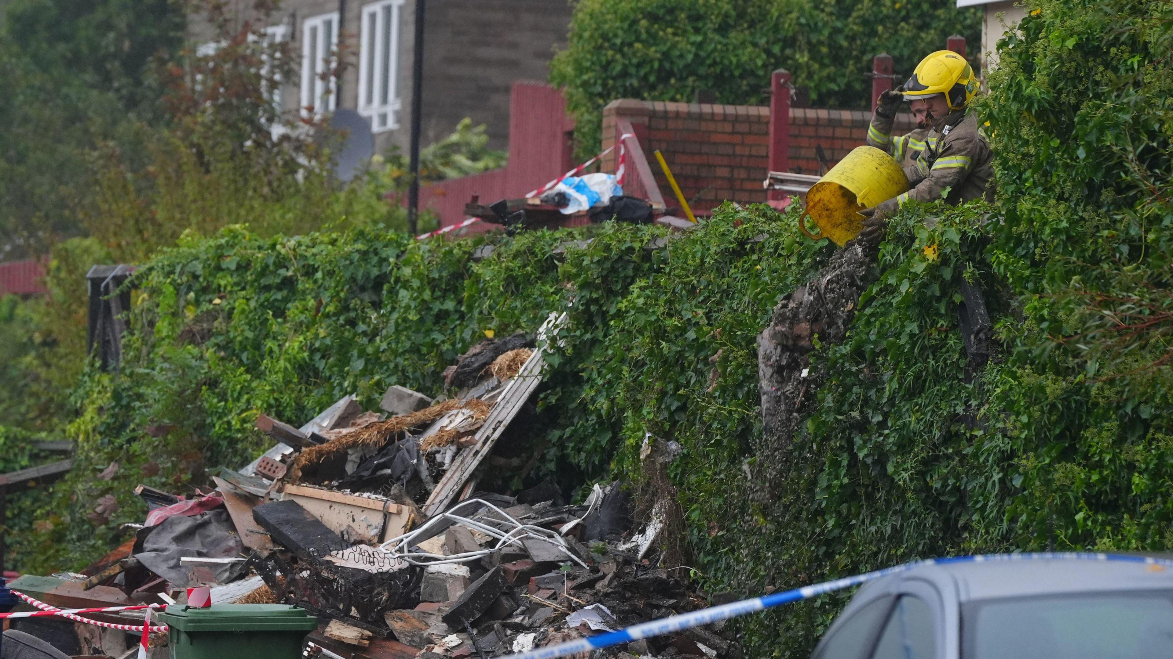 Firefighters appearing to be throwing ash over the side of a hedge into a pile of debris. A police cordon surrounds the scene.